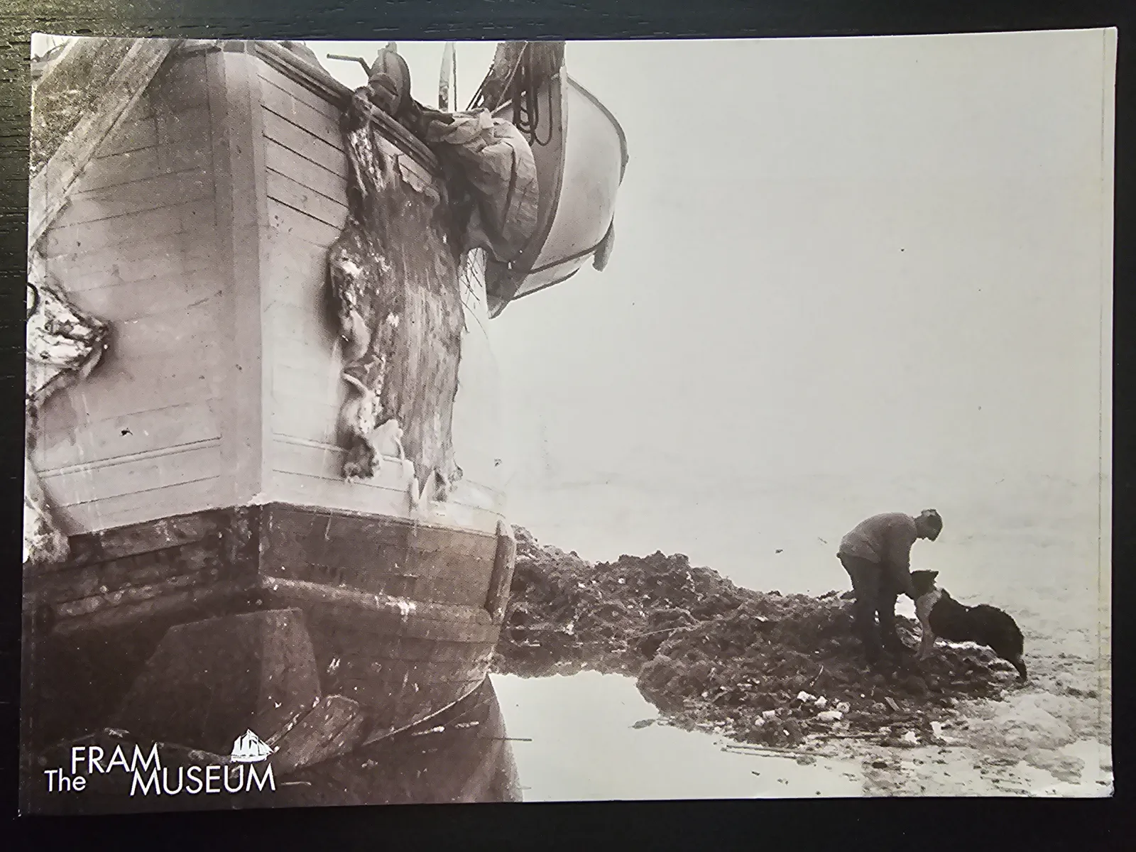 Ein Schwarz-Weiß-Foto zeigt ein Schiff mit hängenden Tierfellen und einen Mann mit Hund auf unebenem Gelände.