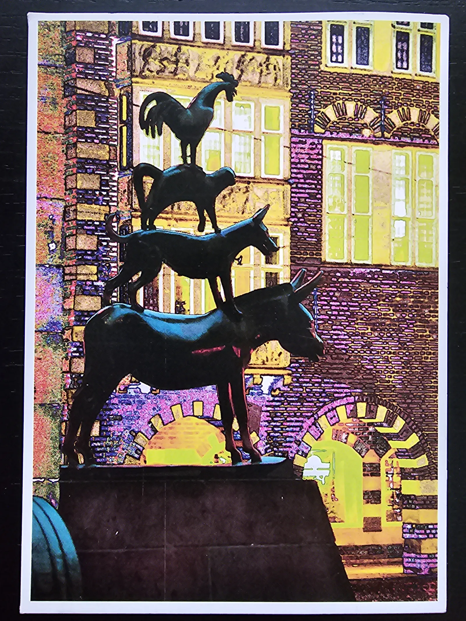 Eine dunkle Statue der Bremer Stadtmusikanten – Esel, Hund, Katze und Hahn übereinander – steht vor einem farbenfroh stilisierten Gebäude.