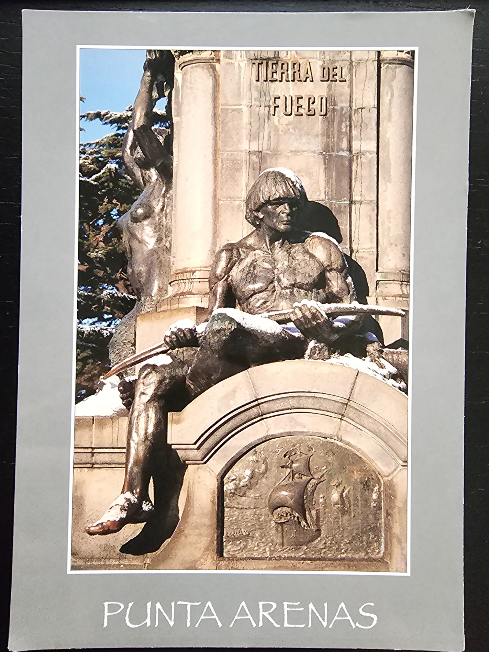 Eine schneebedeckte Bronzestatue eines sitzenden Mannes, Teil eines Denkmals mit der Inschrift "Tierra del Fuego", steht vor einem blauen Himmel in Punta Arenas.