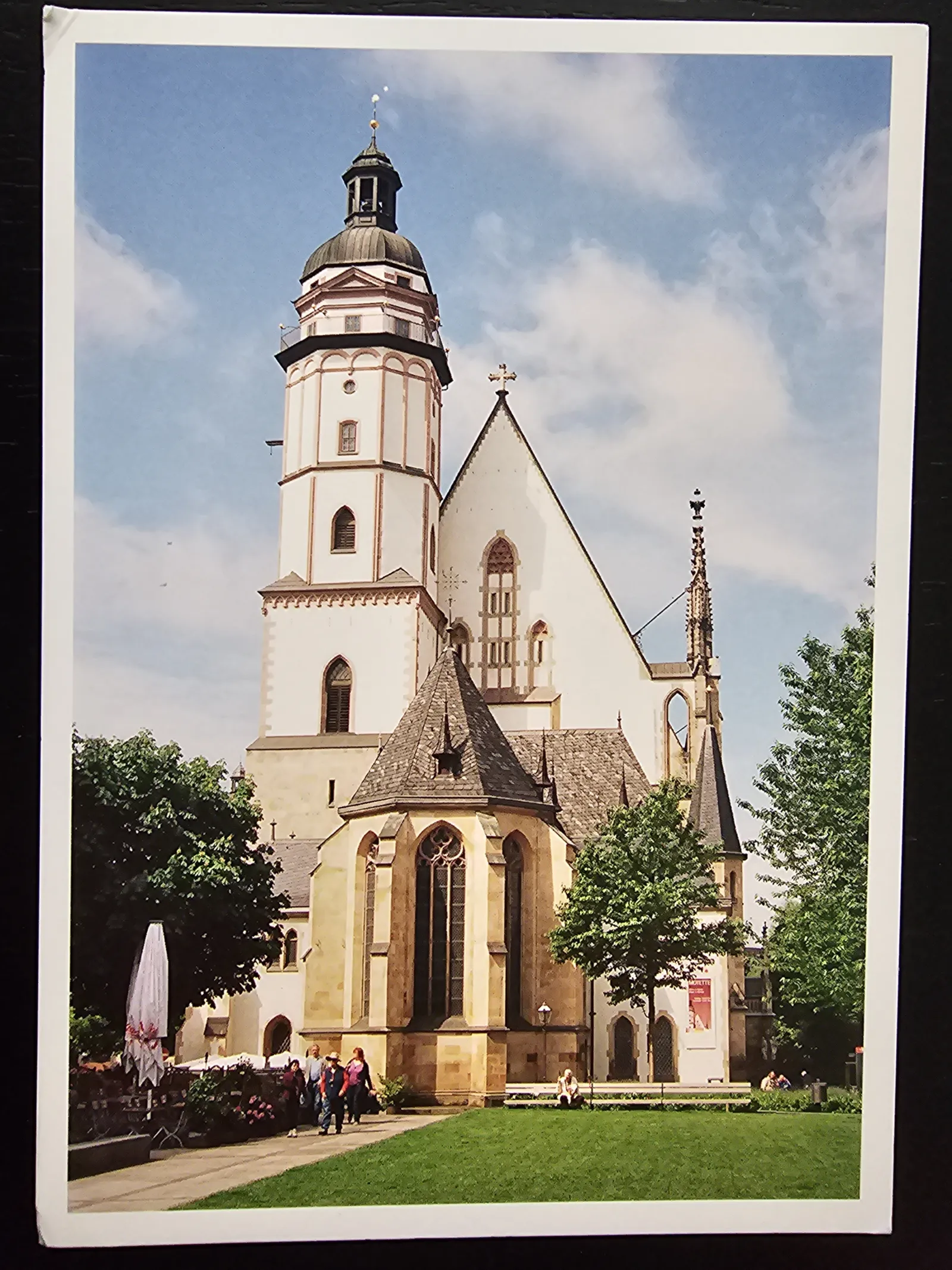 Eine große, helle Kirche mit einem hohen Turm und Kuppel ist von Grünflächen, Bäumen und einigen Menschen unter blauem Himmel umgeben.