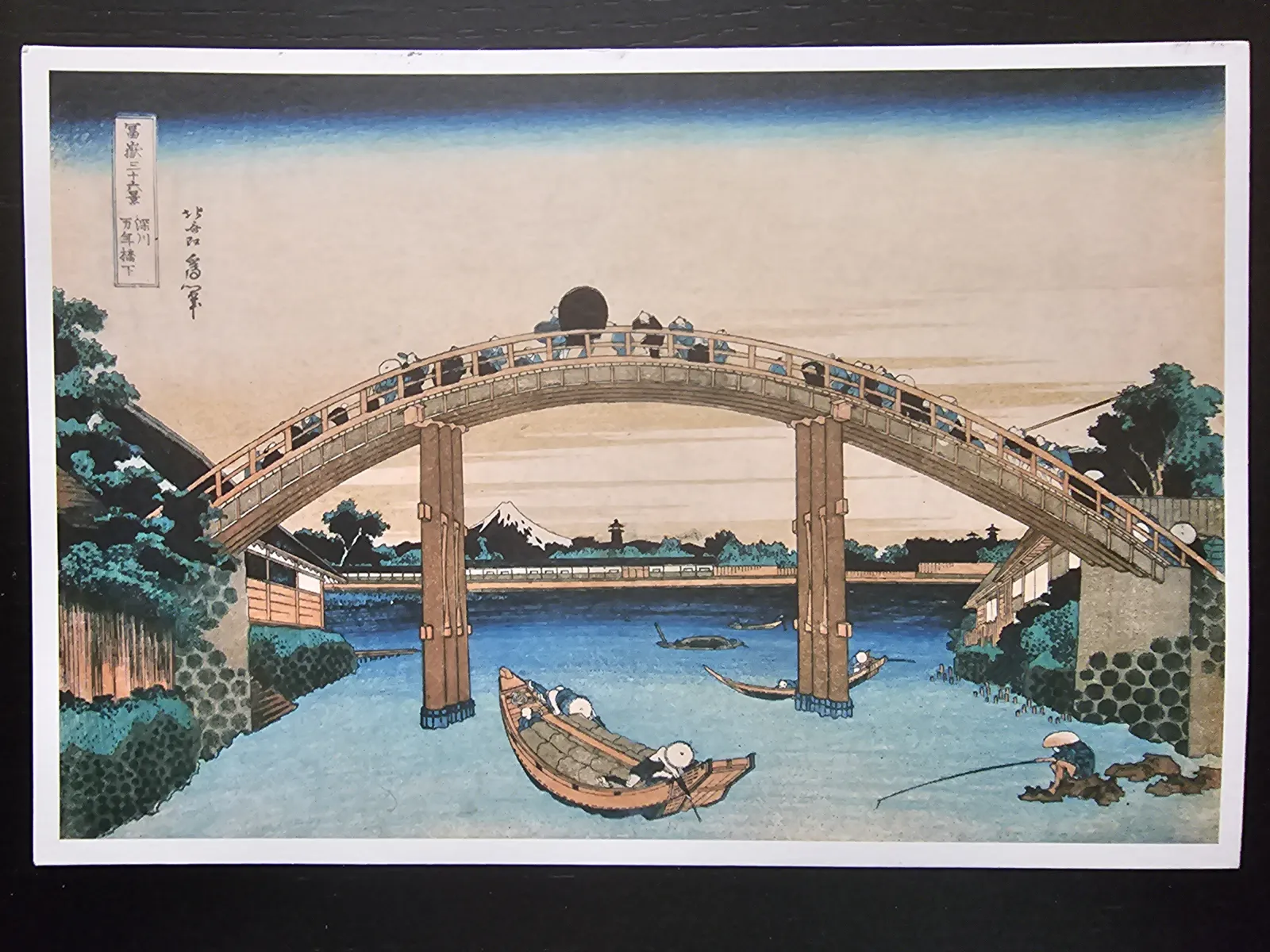 Ein japanischer Farbholzschnitt zeigt eine belebte, bogenförmige Holzbrücke über einem Fluss, mit dem Berg Fuji im Hintergrund.