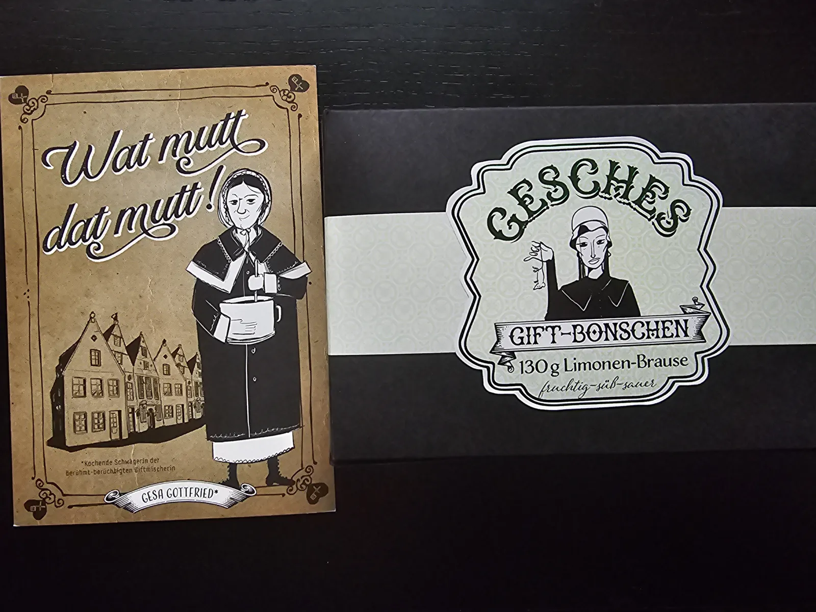 Eine braune Karte mit einer altmodisch gekleideten Frau und dem Namen "Gesa Gottfried" liegt neben einer schwarzen Geschenkbox mit einem Etikett für "Gesches Gift-Bonschen".