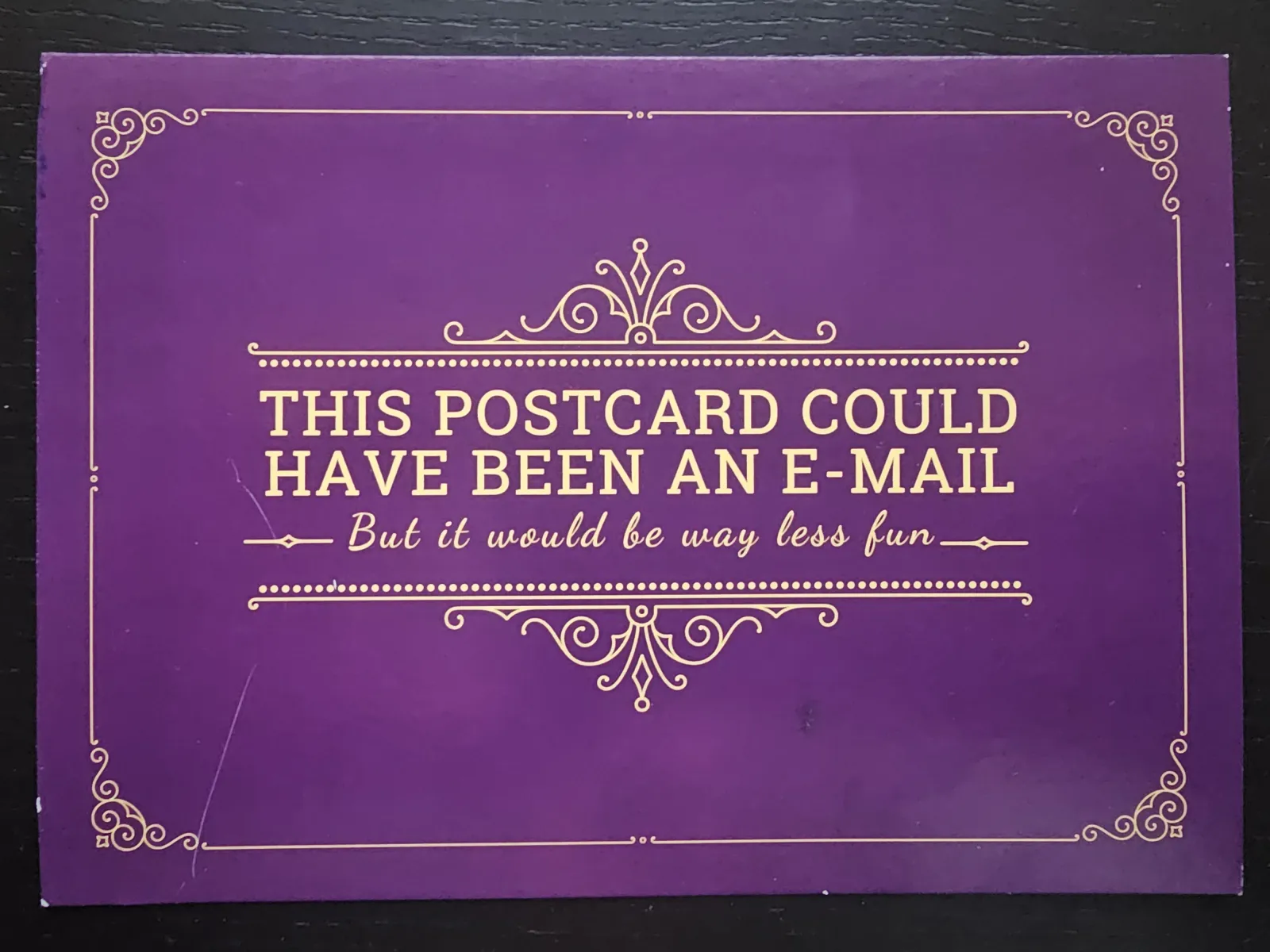 Eine violette Postkarte mit goldenem Zierrahmen und dem englischen Text: „THIS POSTCARD COULD HAVE BEEN AN E-MAIL – But it would be way less fun –“.