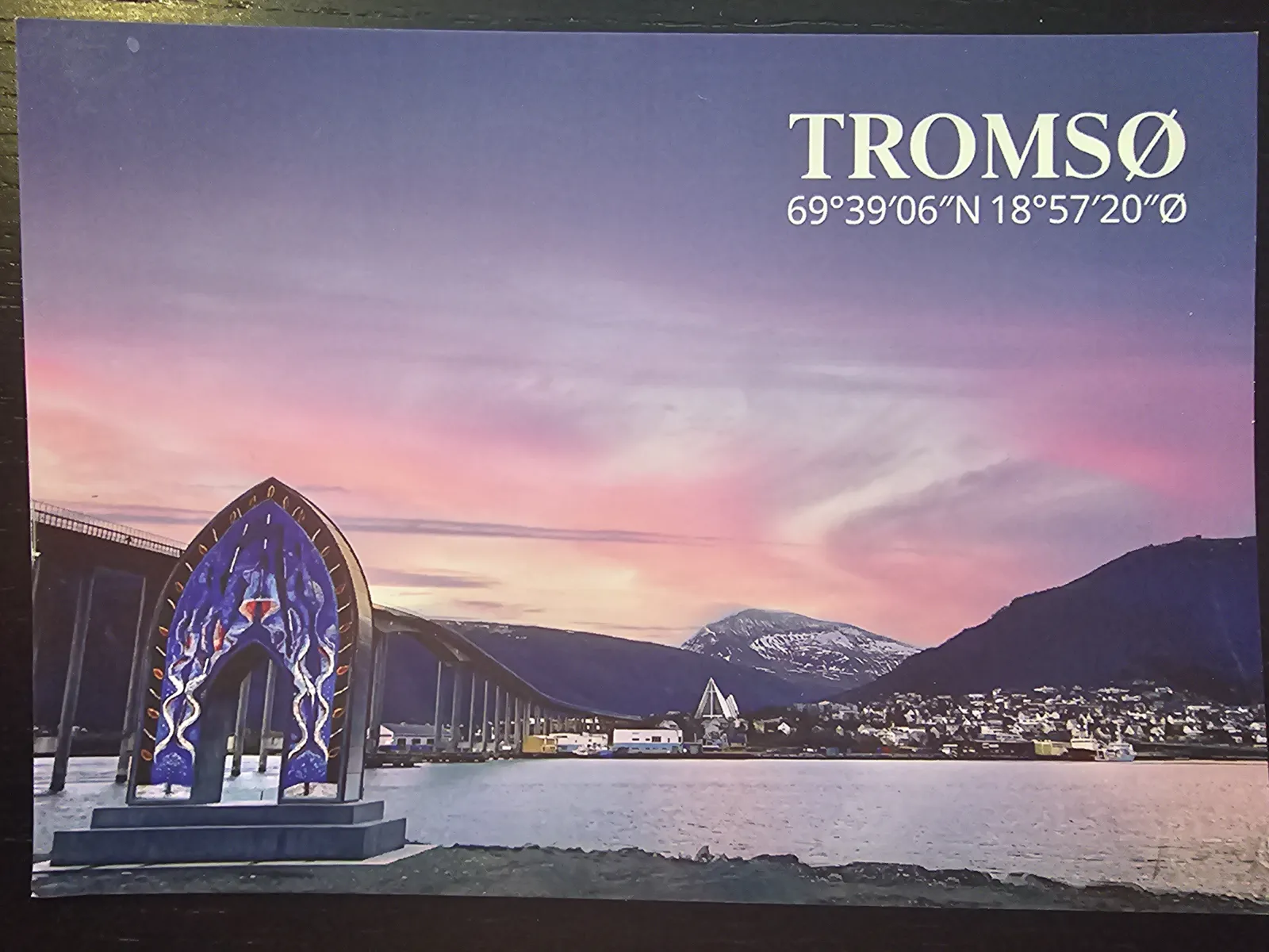 Tromsø bei Sonnenuntergang, mit einem markanten blauen Bogen im Vordergrund, der Stadt am Wasser und Bergen unter einem rosa-lila Himmel.