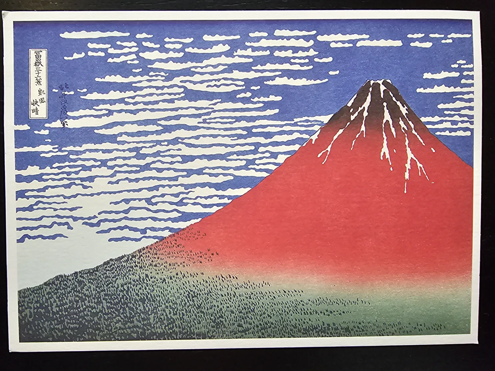 Der japanische Holzschnitt zeigt den rotbraunen Berg Fuji mit weißen Schneeflecken, einem blauen Himmel mit weißen Wolken und grünen Wäldern am Fuße.