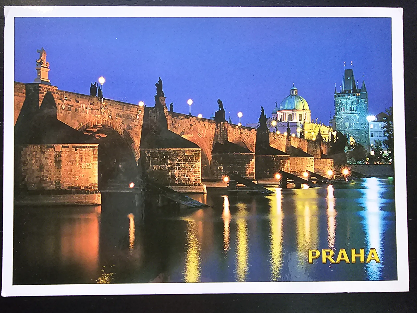 Die beleuchtete Karlsbrücke in Prag ist nachts mit Statuen und ihren Spiegelungen im Fluss zu sehen, vor einer dunklen Stadtsilhouette mit Kuppel und Turm.