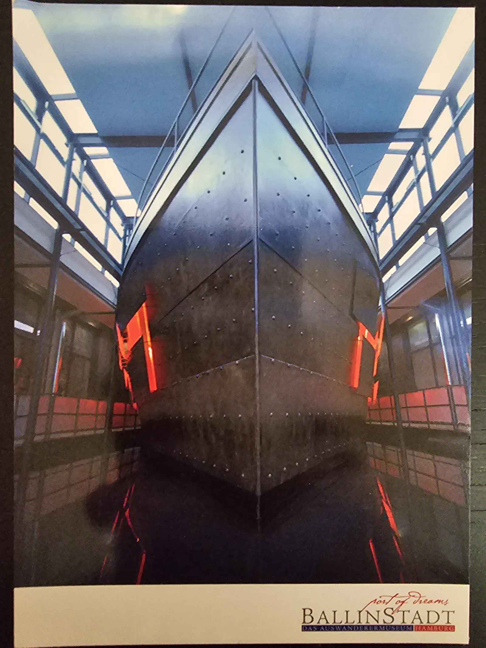 Ein dunkler, genieteter Schiffsbug ragt symmetrisch von unten in die Höhe, umgeben von einer hellblauen Deckenkonstruktion mit weißen Balken und roten Lichtakzenten.