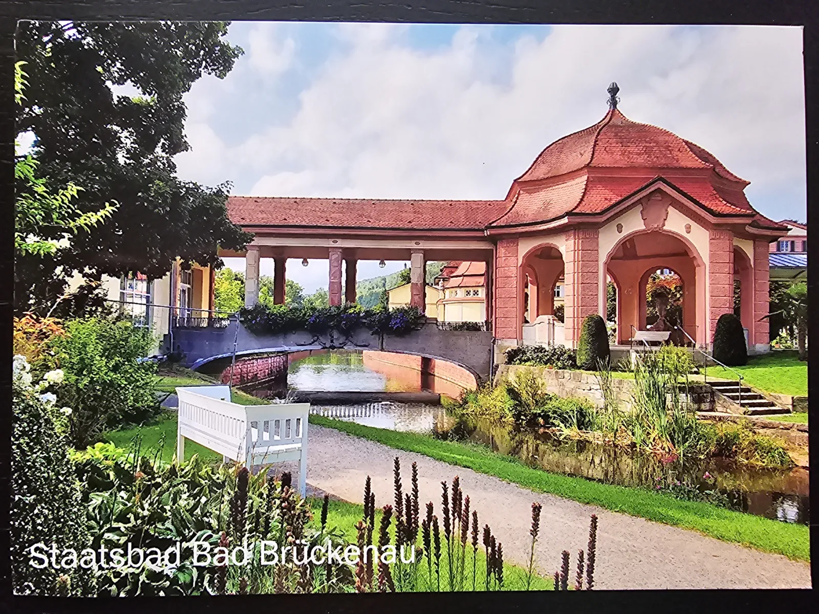 Roter Pavillon und säulengestützte Brücke überspannen einen Bach in einer grünen Parkanlage des Staatsbades Bad Brückenau.