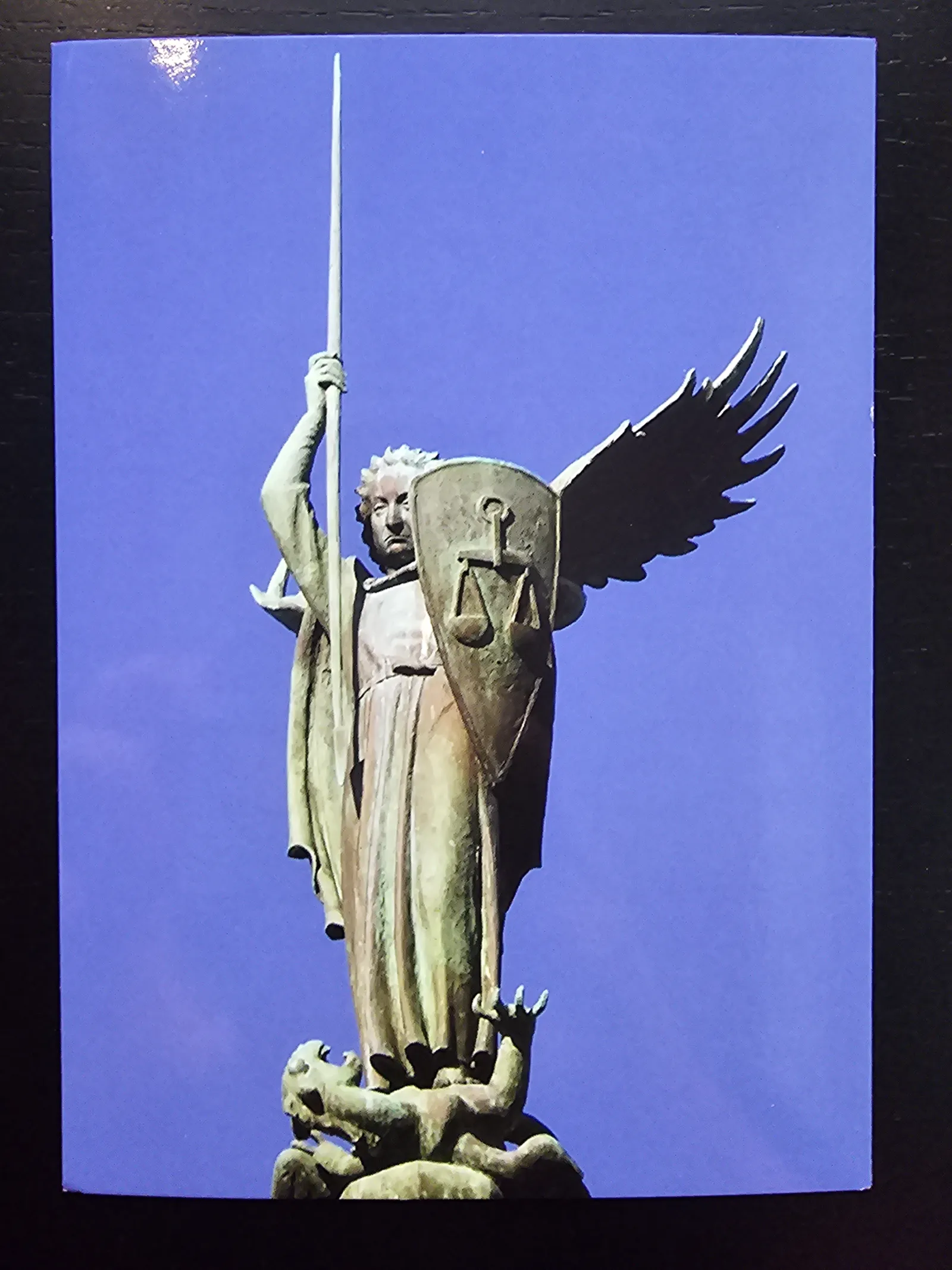 Eine bronzene Statue eines geflügelten Engels mit Speer und Schild, der auf einem besiegten Drachen steht, vor blauem Himmel.