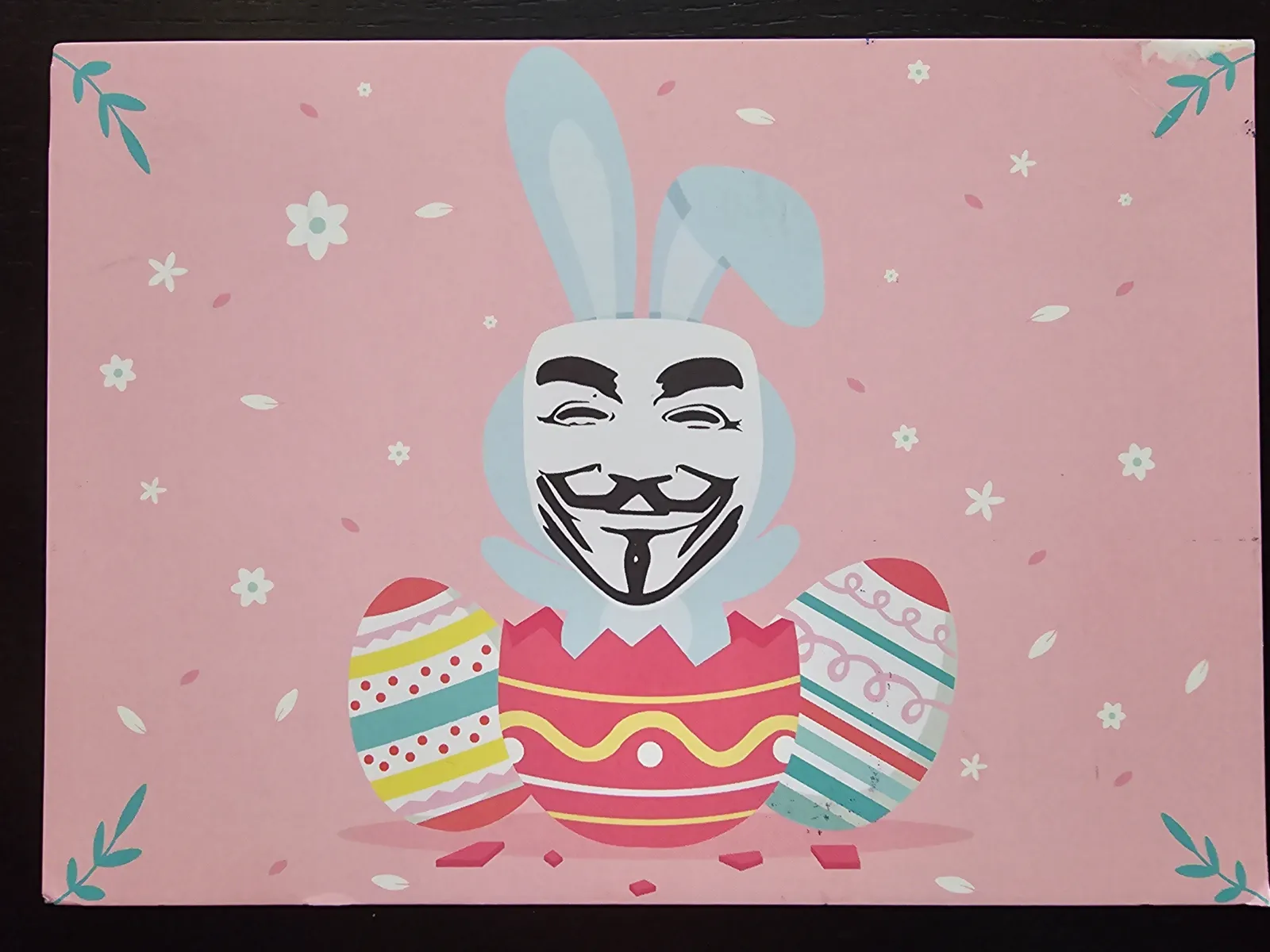 Auf rosafarbenem Hintergrund schlüpft ein Osterhase mit Guy-Fawkes-Maske aus einem zerbrochenen Osterei, flankiert von zwei weiteren bunten Eiern und kleinen Blumen.