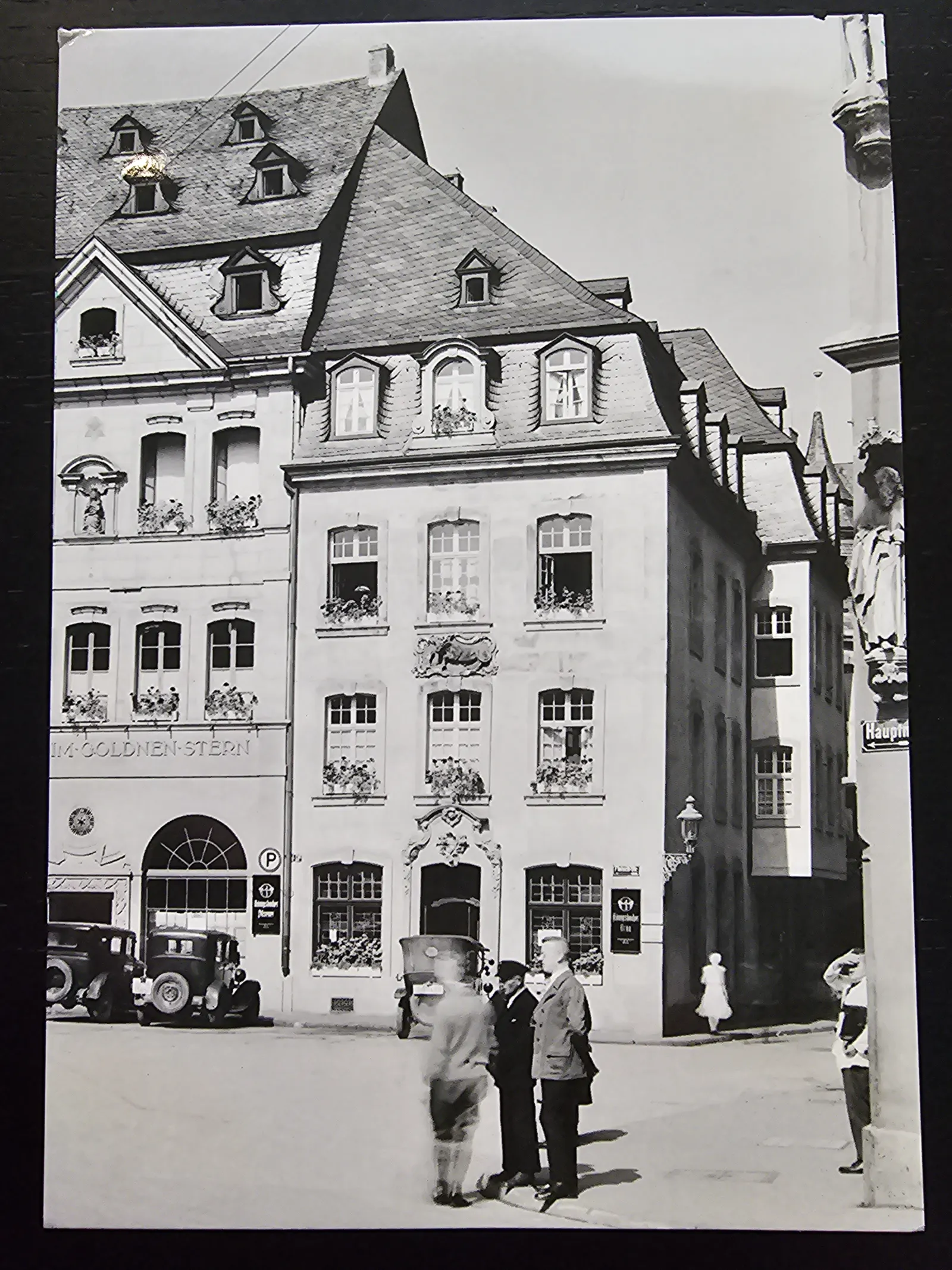 Ein Schwarz-Weiß-Foto zeigt eine belebte historische Straße mit mehrstöckigen Gebäuden, geparkten Oldtimern und mehreren Personen.