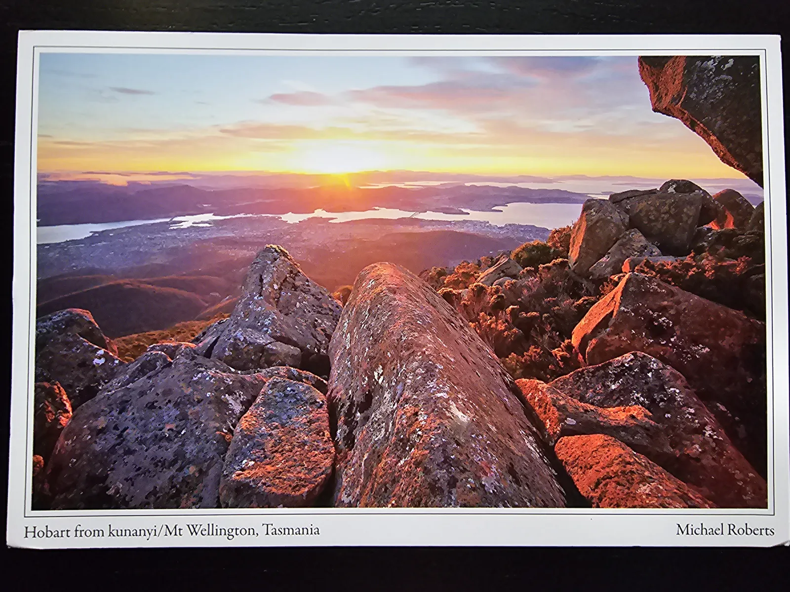 Eine weite Ansicht von einem felsigen Berg bei Sonnenuntergang, mit einer Stadt und einem Gewässer in der Ferne.