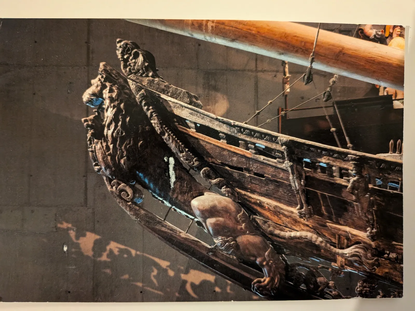 Ein Nahaufnahme des Bugs eines alten, dunklen Holzschiffs mit aufwendigen Schnitzereien und einer großen löwenähnlichen Figur vor dunklem Hintergrund.