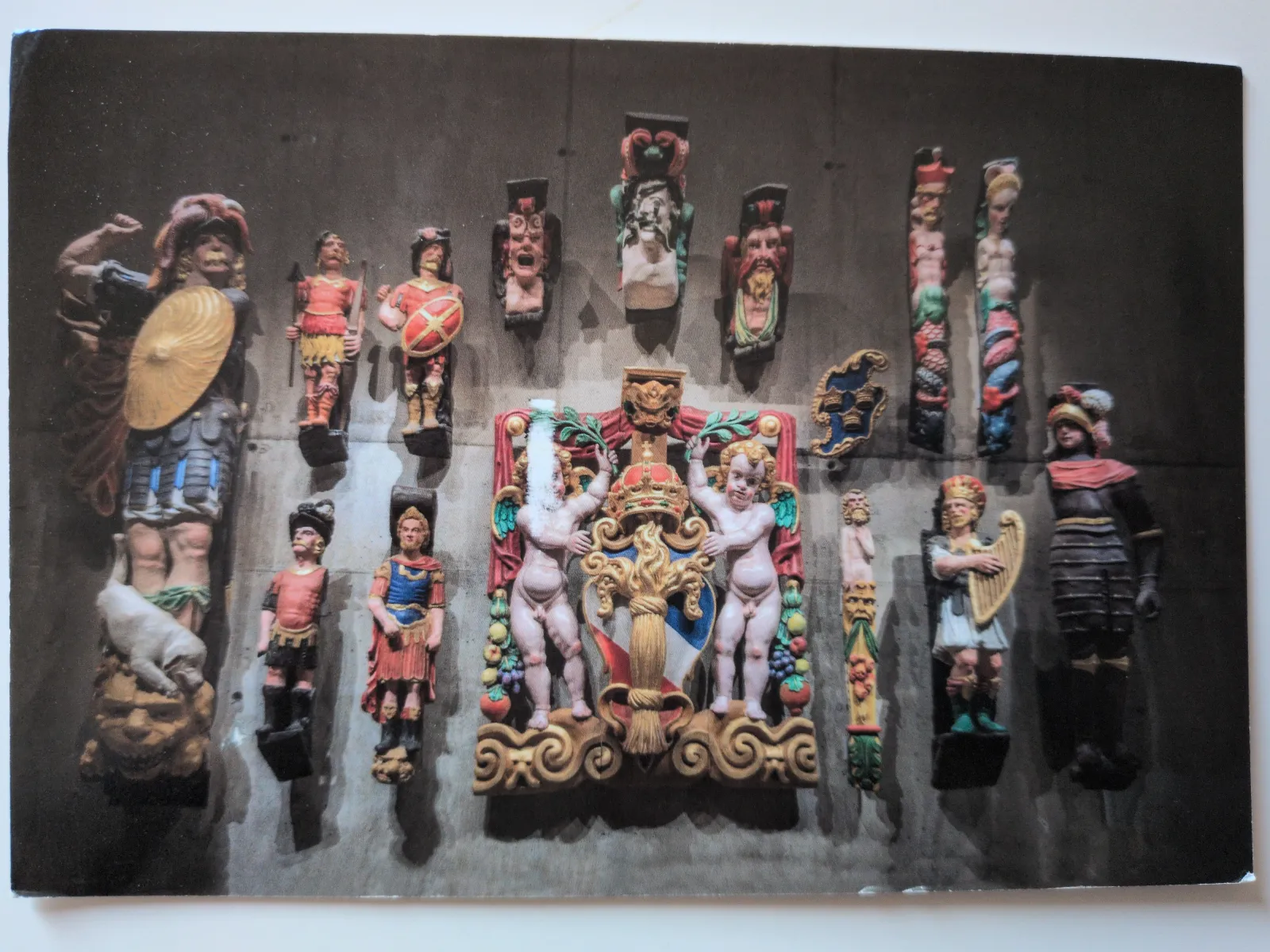 Eine Anordnung von farbig bemalten, geschnitzten Holzfiguren, darunter Krieger und Putten, ist auf einer dunklen Wand montiert.