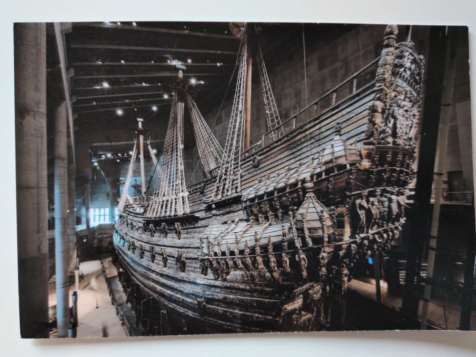Ein riesiges, dunkles Holzschiff mit hohen Masten und kunstvollen Schnitzereien am Heck steht in einem großen, schwach beleuchteten Museumssaal.