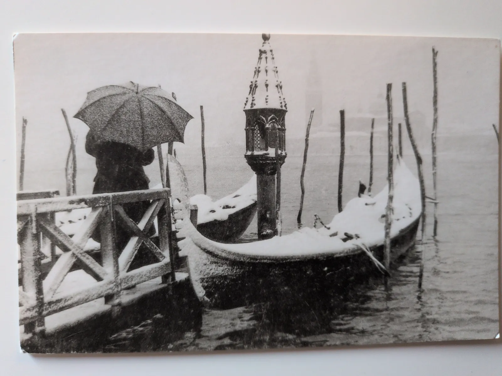 Ein Schwarz-Weiß-Bild zeigt schneebedeckte Gondeln, eine Person mit Regenschirm und einen verzierten Laternenpfahl in einem winterlichen Venedig.
