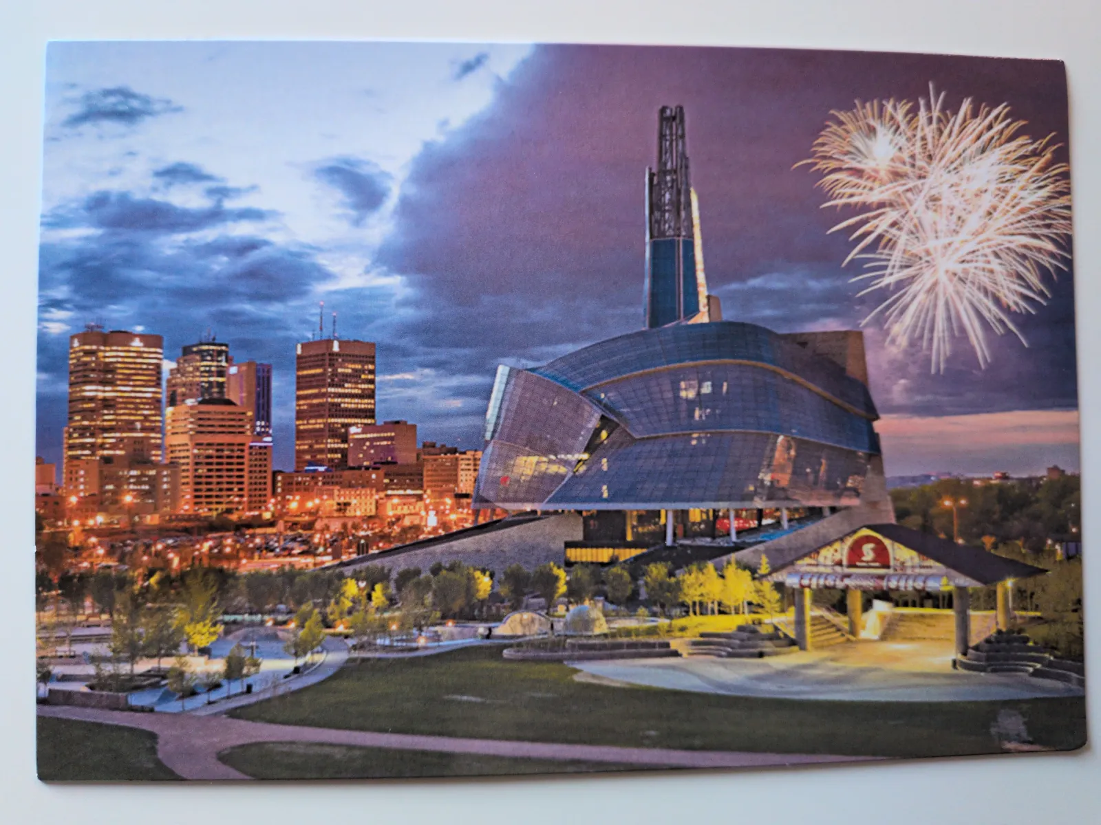 Ein markantes, geschwungenes Gebäude mit hohem Turm dominiert eine nächtliche Stadtlandschaft mit beleuchteter Skyline und Feuerwerk am Nachthimmel.