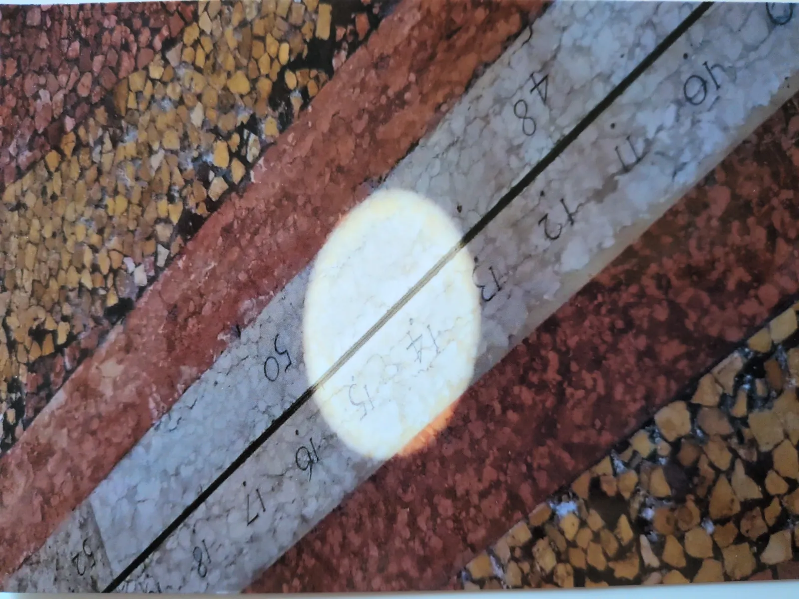 Eine steinerne Sonnenuhr mit einer hellen, nummerierten Skala, einem dunklen Zeiger und einem leuchtenden Lichtfleck ist auf einem Mosaikuntergrund abgebildet.