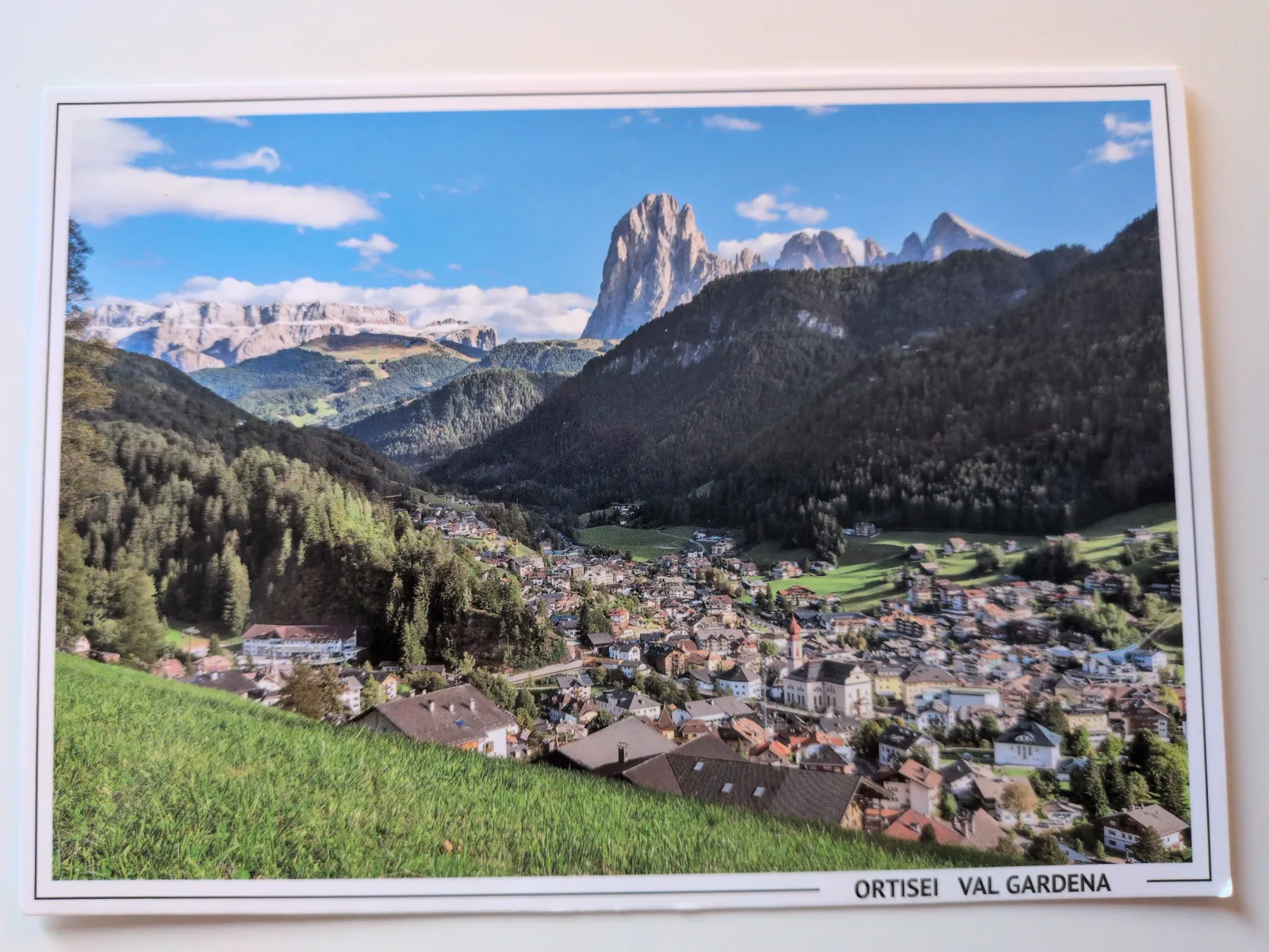Blick auf das Bergdorf Ortisei im grünen Tal, umrahmt von bewaldeten Hängen und den imposanten Dolomiten unter blauem Himmel.