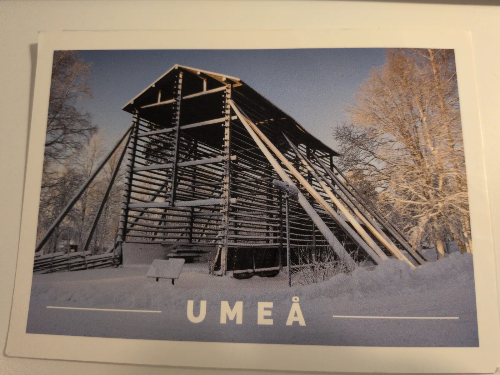 Ein hohes, offenes Holzgestell steht in einer tief verschneiten Winterlandschaft mit dem Schriftzug UMEÅ am unteren Bildrand.