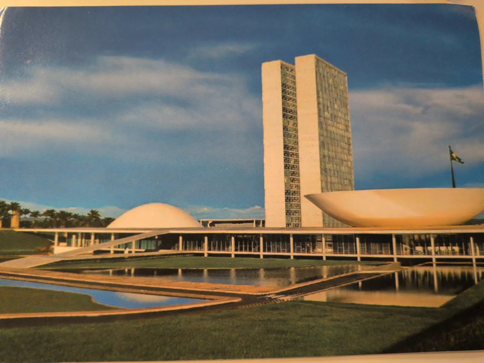 Das Bild zeigt den modernen Nationalkongress Brasiliens mit zwei hohen Türmen, einer Kuppel und einer Schale, umgeben von Wasserbecken und Grünflächen unter blauem Himmel.