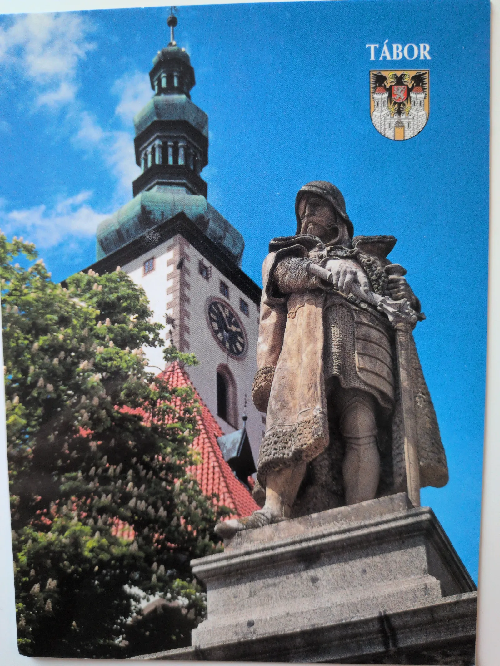 Eine steinerne Kriegerstatue steht vor einem hohen Kirchturm mit Uhr und grünen Bäumen unter blauem Himmel in Tábor.