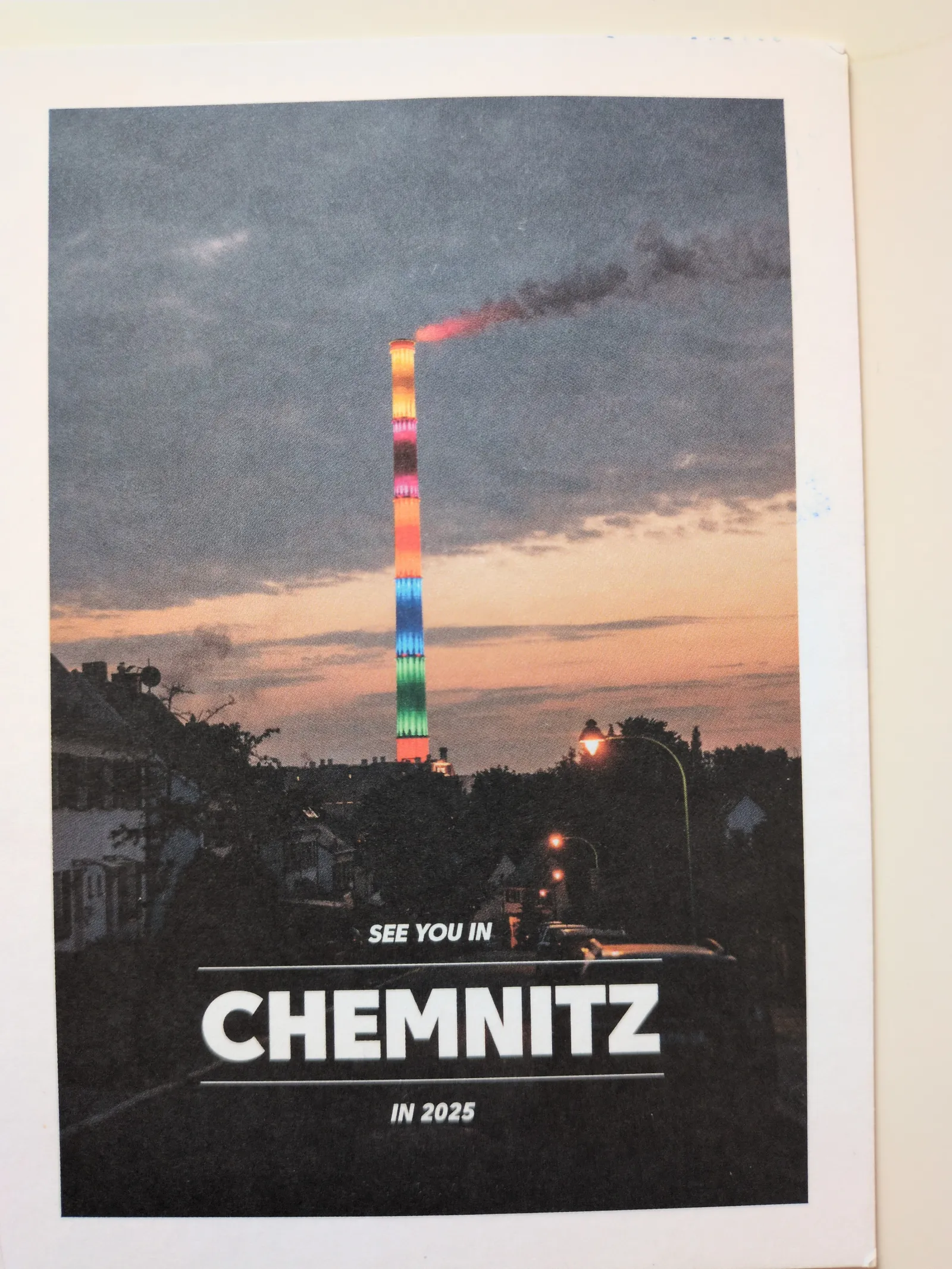 Ein hoher Schornstein leuchtet bei Dämmerung in bunten Regenbogenfarben über einer Stadt, darunter steht "SEE YOU IN CHEMNITZ IN 2025".