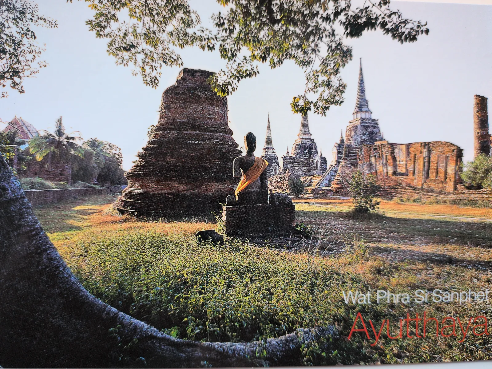 Das Bild zeigt die sonnigen Ruinen des historischen Tempels Wat Phra Si Sanphet in Ayutthaya mit einer sitzenden Buddha-Statue im Vordergrund, umgeben von mehreren spitzen Stupas und alten Ziegelstrukturen inmitten von Gras und Bäumen.