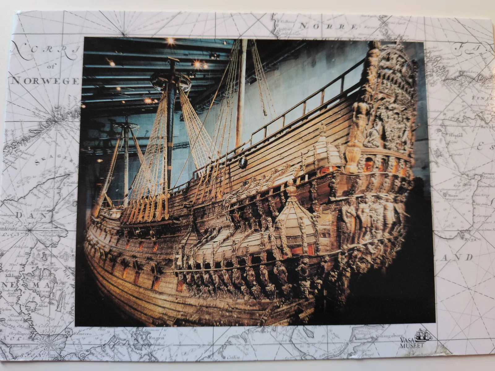 Ein großes, reich verziertes Holzschiff mit Masten ist im Vasa Museum ausgestellt, umrahmt von einer historischen Seekarte.