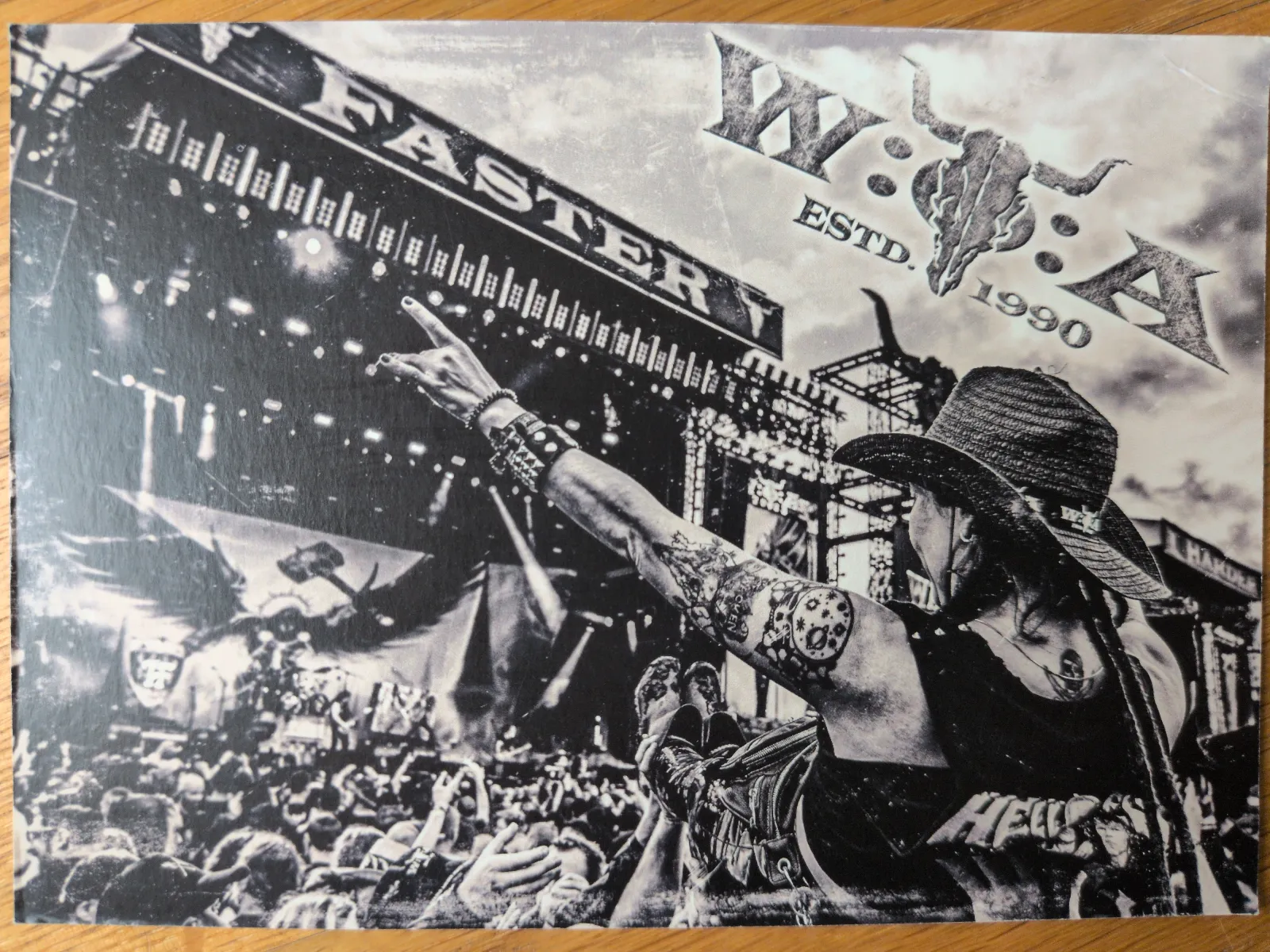 Schwarz-Weiß-Foto einer Person mit Strohhut und tätowierten Armen, die bei einem Wacken-Konzert die Pommesgabel in Richtung Bühne und jubelnder Menge macht.