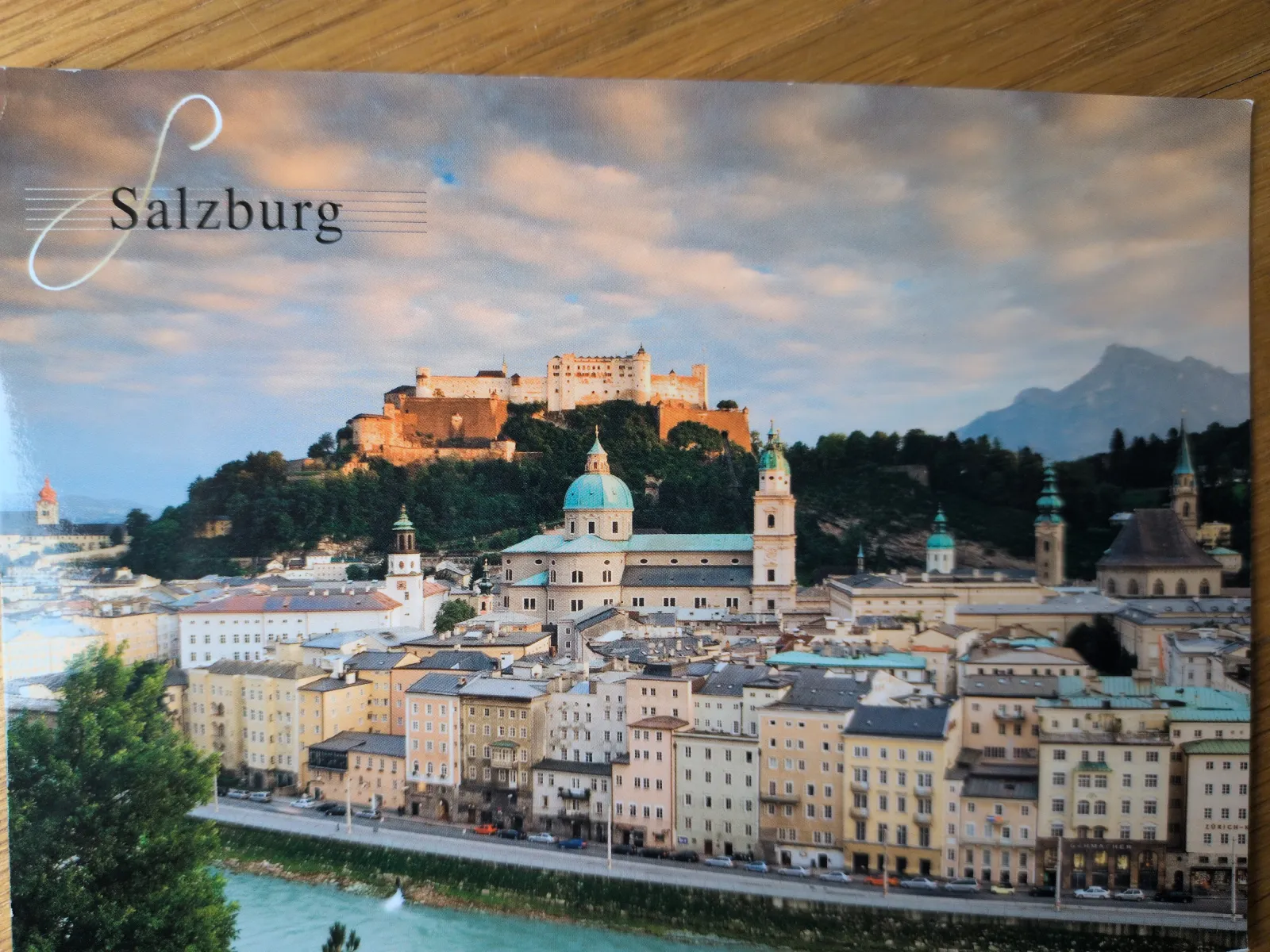 Das Bild zeigt Salzburg in warmem Licht unter bewölktem Himmel, mit der Festung Hohensalzburg auf einem Hügel, dem Dom mit seinen Kuppeln, der Altstadt am Fluss Salzach und Bergen im Hintergrund.