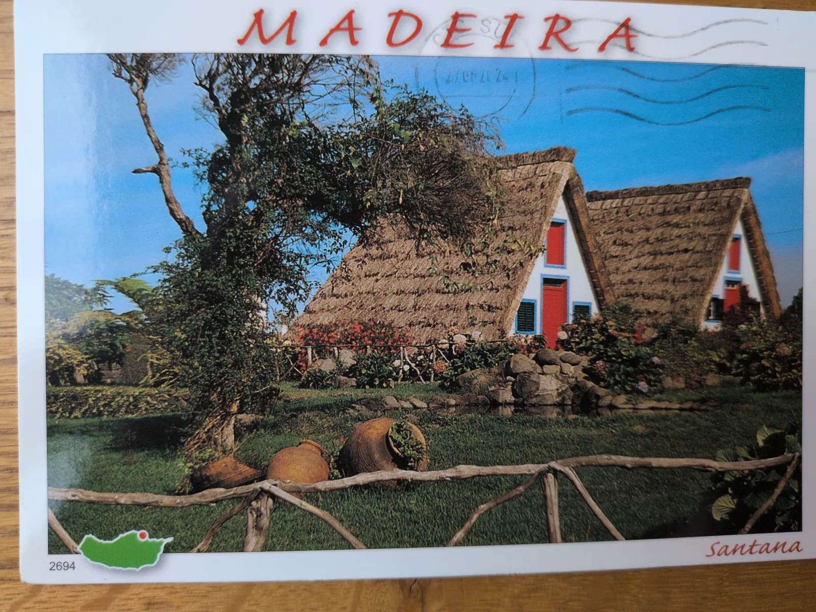 Zwei traditionelle Häuser mit hohen Strohdächern und roten Türen stehen in einer grünen, blühenden Landschaft.