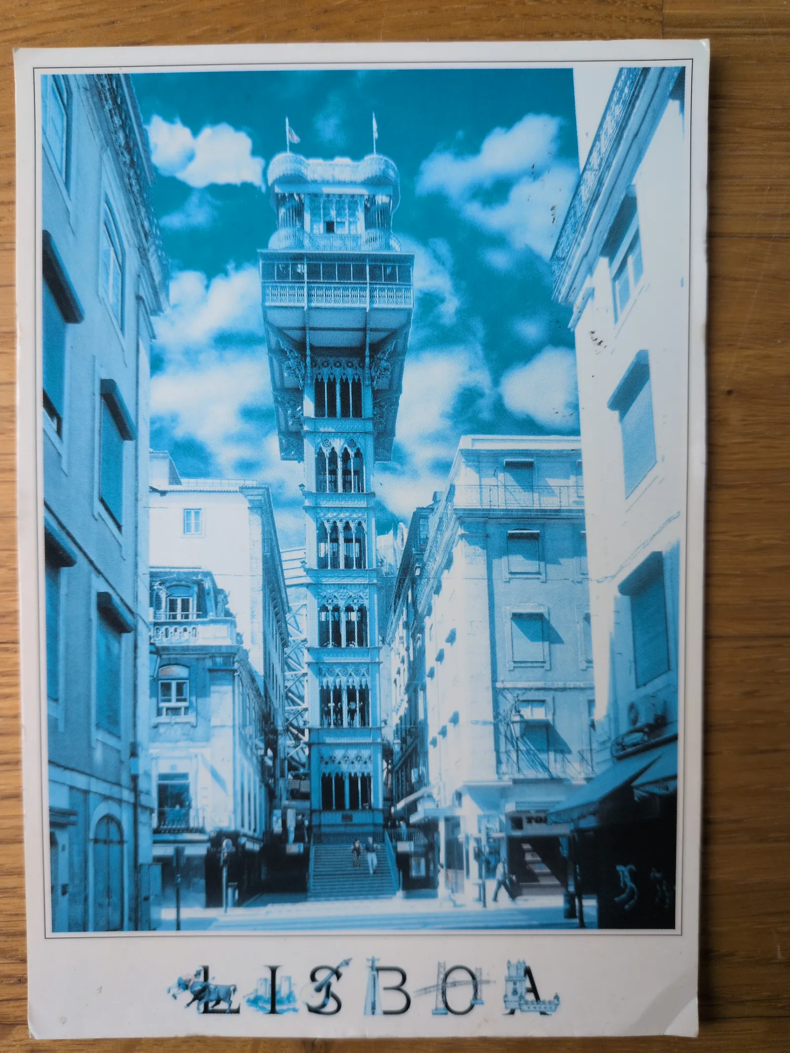 Eine blau getönte Postkarte zeigt den hohen, verzierten Elevador de Santa Justa in Lissabon, umgeben von Stadtgebäuden, mit dem Schriftzug „LISBOA“ am unteren Rand.