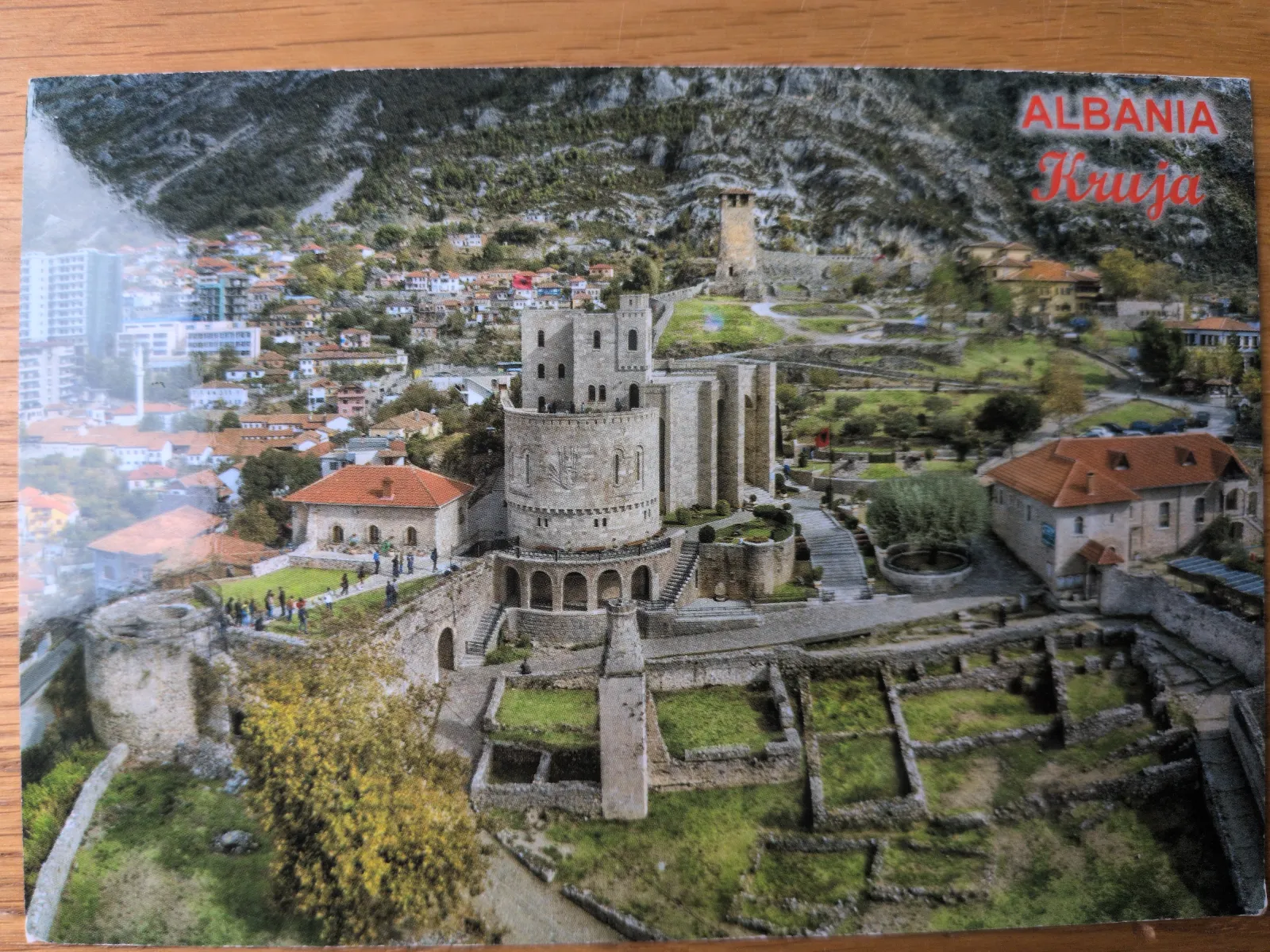 Eine Luftaufnahme zeigt die historische Burg Kruja in Albanien auf einem Hügel über einer Stadt, umgeben von Bergen.