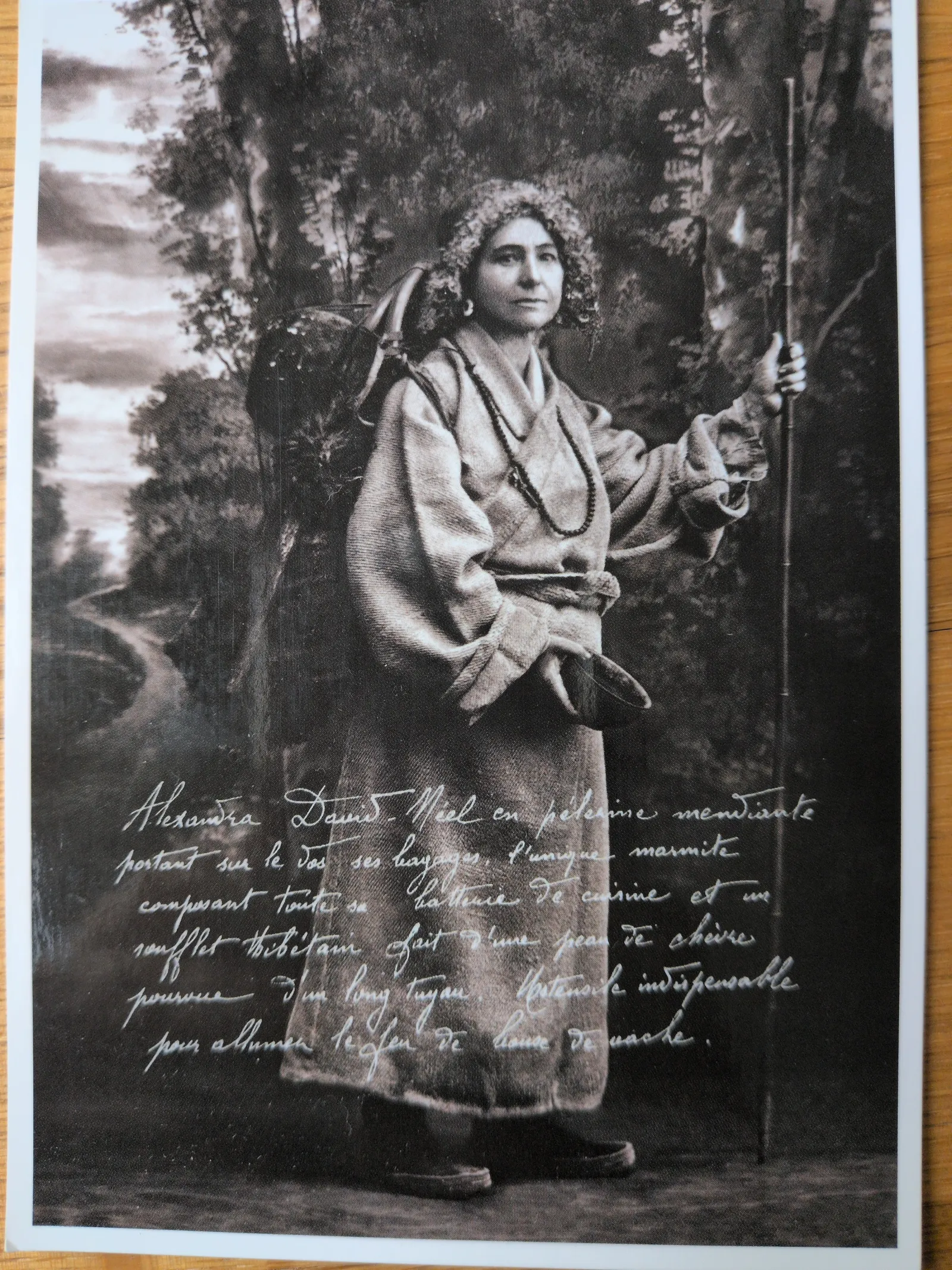 Ein Schwarz-Weiß-Foto zeigt Alexandra David-Néel als Pilgerin mit lockigem Haar, die einen Rucksack auf dem Rücken und einen Wanderstab in der Hand hält.