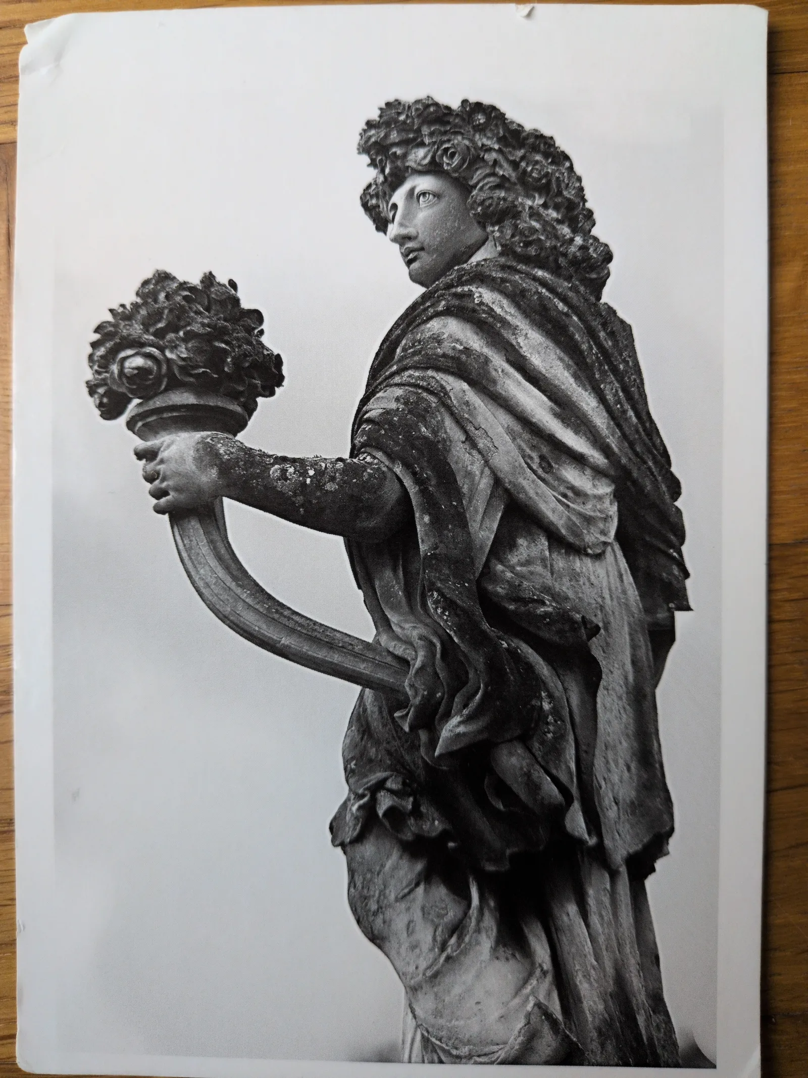 Eine verwitterte, schwarz-weiße Steinstatue zeigt eine Figur mit reicher Lockenpracht, die einen verzierten Kelch mit Blumen und kleinen Gesichtern in der Hand hält und nach links oben blickt.