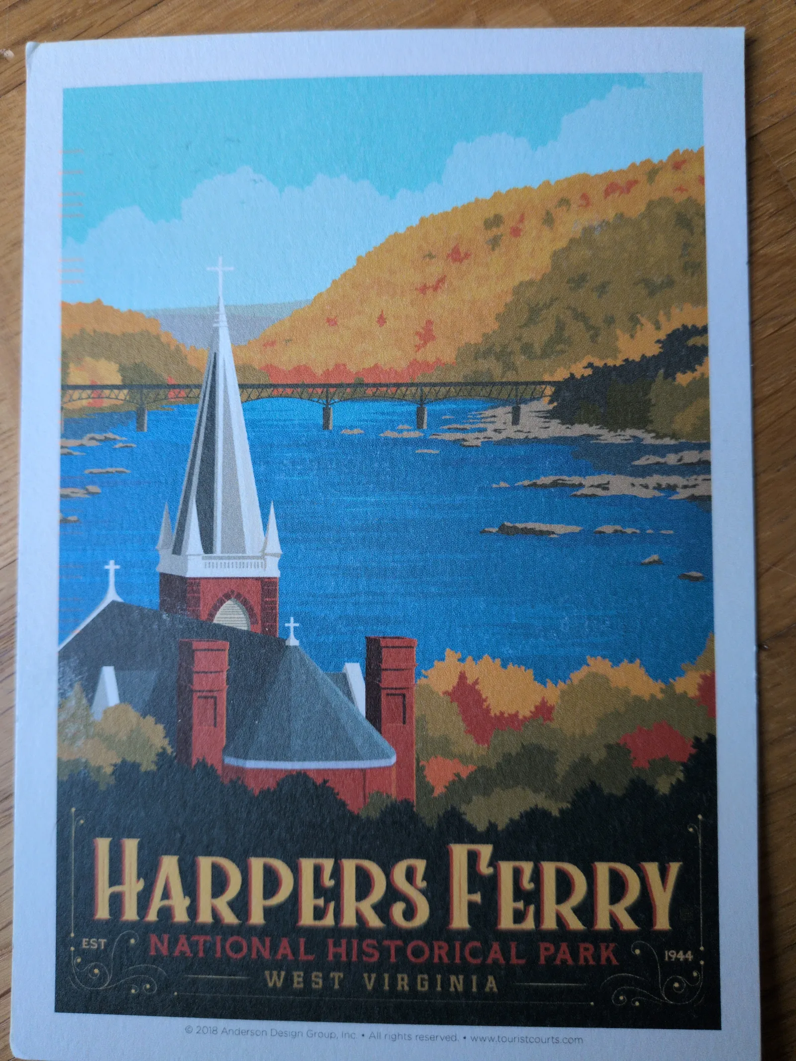 Ein Vintage-Poster zeigt eine Kirche mit spitzem Turm im Vordergrund, dahinter einen Fluss mit Brücke und herbstlich gefärbten Bäumen, sowie den Schriftzug "Harpers Ferry National Historical Park".