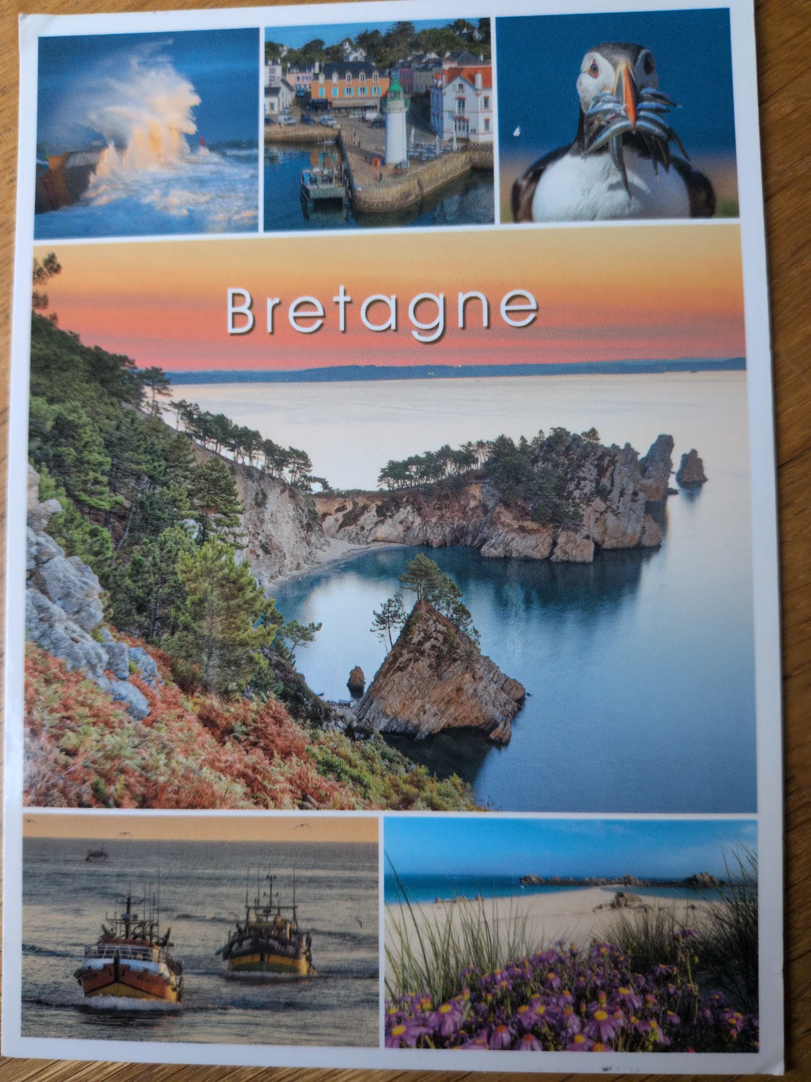 Eine Collage mit dem vertikalen Schriftzug „Bretagne“ zeigt eine große Küstenlandschaft mit Klippen und Strand bei Sonnenuntergang, umgeben von kleineren Bildern wie einem Fischerboot, einem Hafen und einem Papageientaucher.