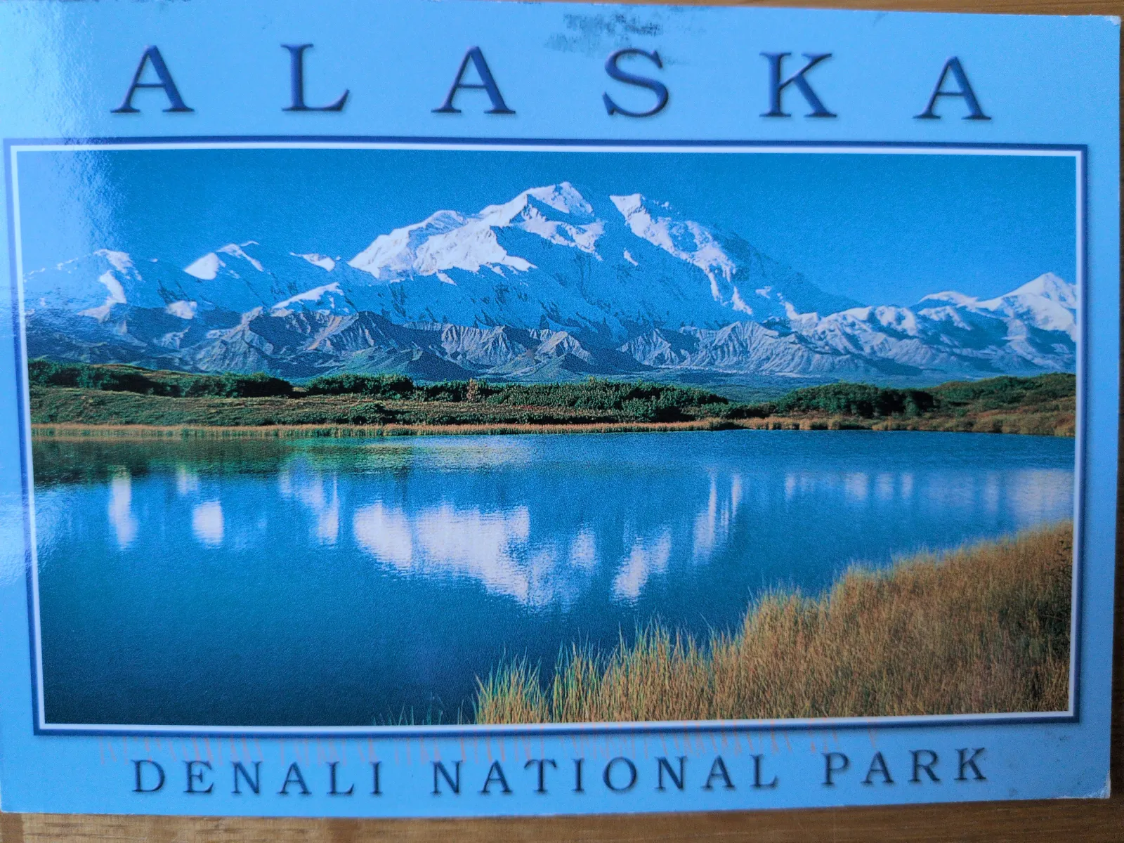 Ein Landschaftsbild des Denali-Nationalparks zeigt einen blauen See, der schneebedeckte Berge und den Himmel widerspiegelt, mit den Titeln 'ALASKA' oben und 'DENALI NATIONAL PARK' unten.