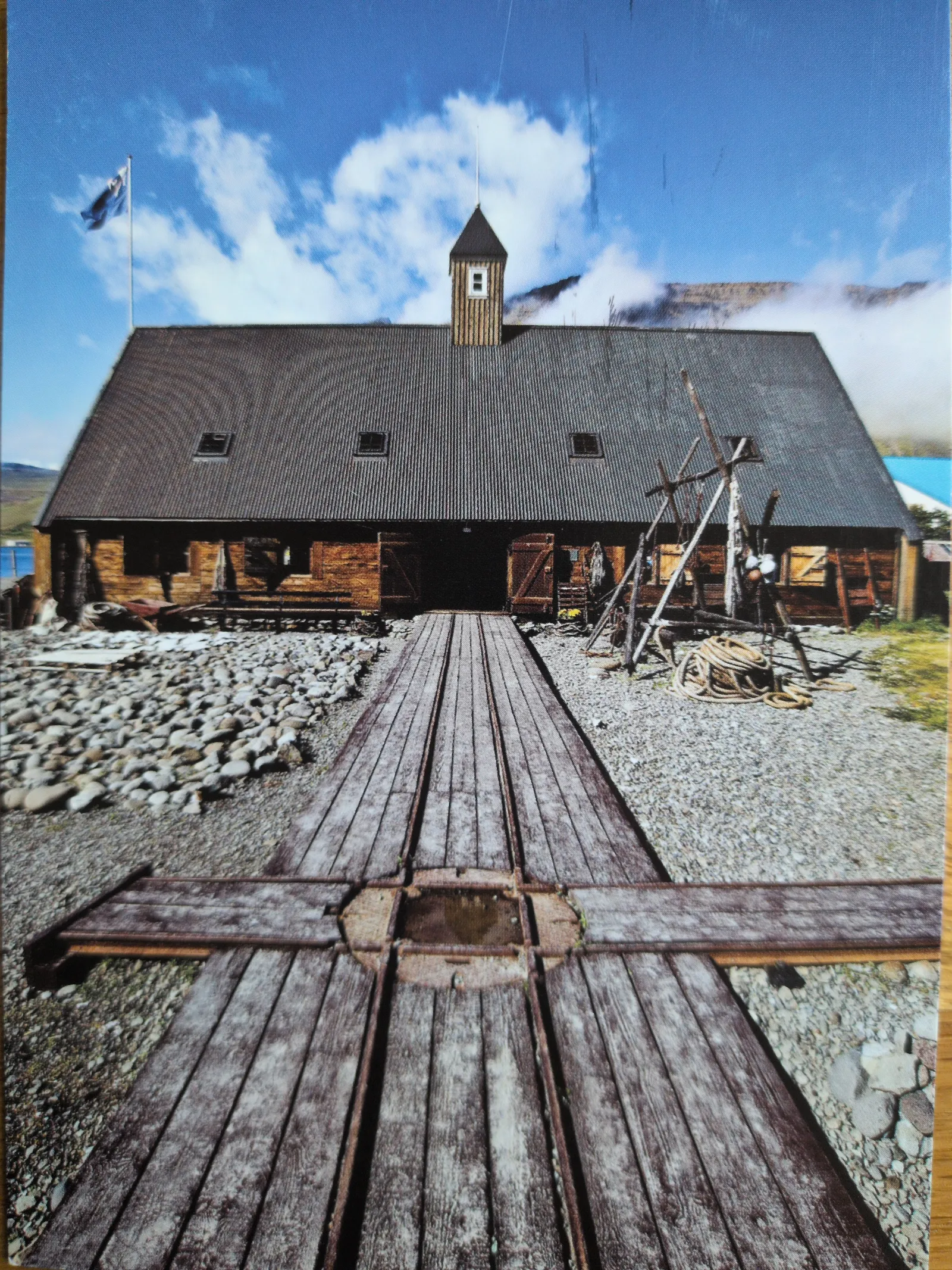 Ein großes, altes Holzgebäude mit einem Bootslip und einer wehenden Flagge steht vor einer Berglandschaft unter blauem Himmel.