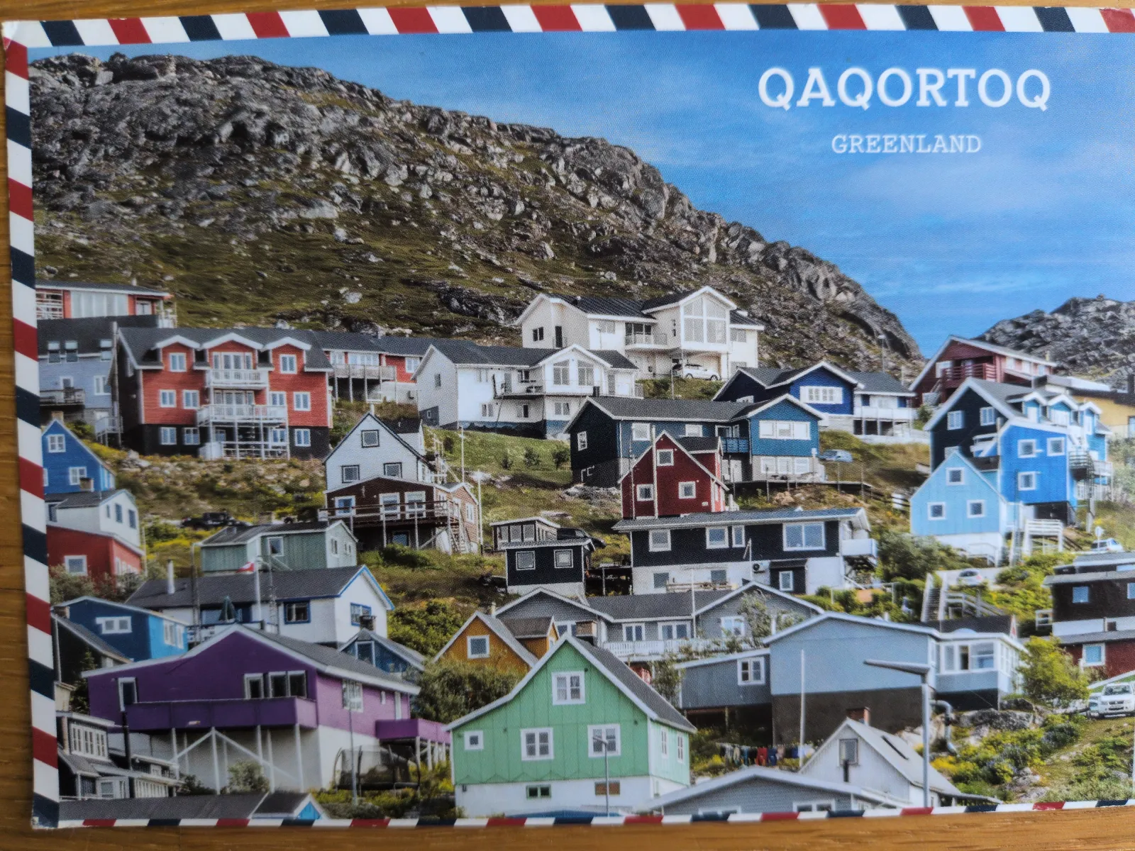 Ein farbenfrohes Dorf mit vielen Häusern schmiegt sich an einen felsigen, grünen Berghang unter blauem Himmel; oben rechts steht "QAQORTOQ GREENLAND".