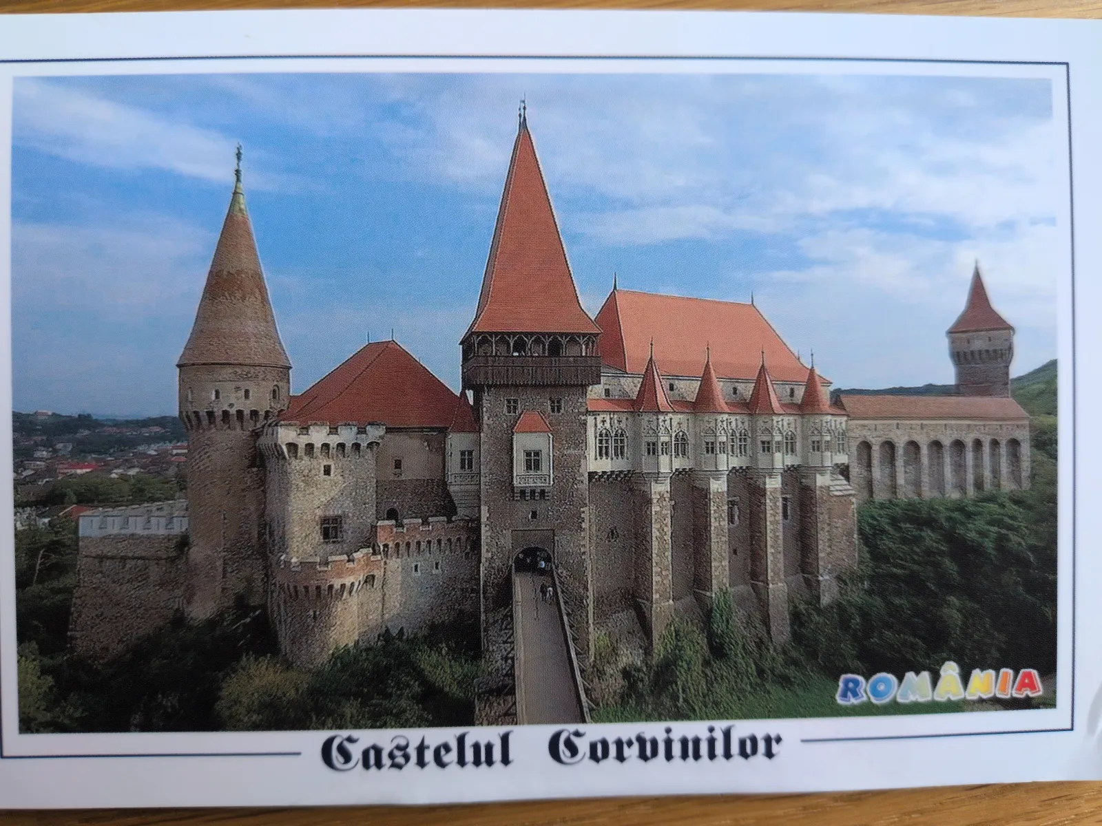 Ein imposantes, mittelalterliches Schloss mit zahlreichen Türmen und roten Dächern, verbunden durch eine lange Brücke, thront in grüner Landschaft.