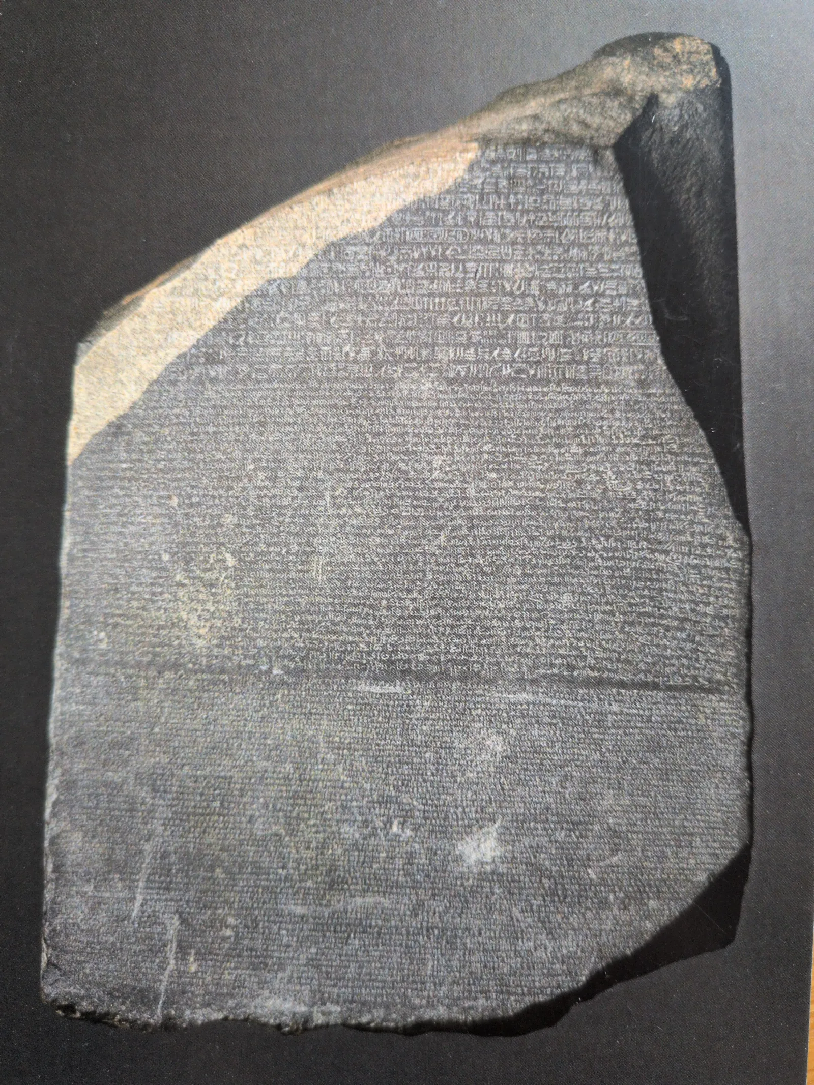 Eine dunkle, unregelmäßig geformte Steinplatte ist dicht mit verschiedenen antiken Schriften bedeckt. Es handelt sich um den Rosetta Stone.