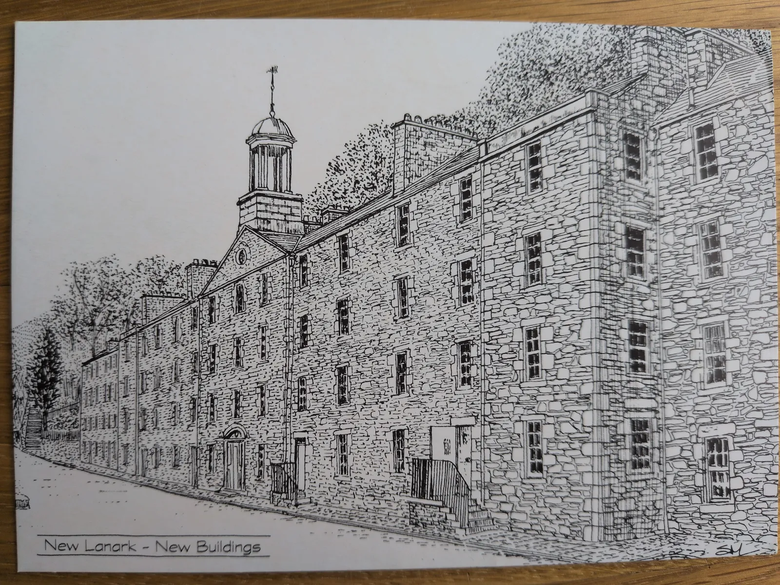 Eine Schwarz-Weiß-Zeichnung zeigt die langen Steingebäude von New Lanark mit zahlreichen Fenstern, einer zentralen Kuppel und Bäumen entlang einer Straße.