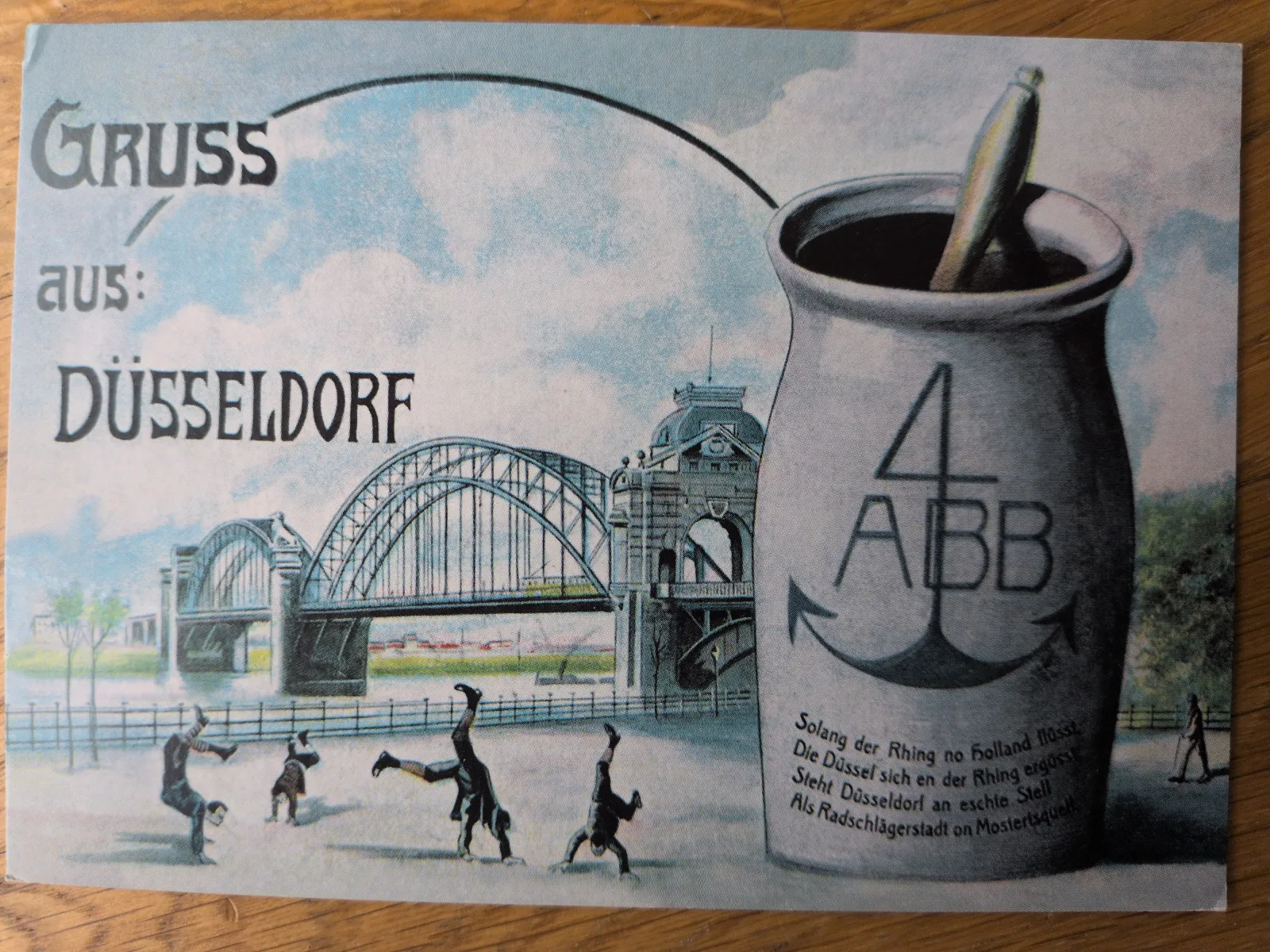 Eine alte Postkarte aus Düsseldorf zeigt eine Rheinbrücke, Radschläger und einen großen Senftopf mit der Aufschrift „Gruss aus Düsseldorf“.