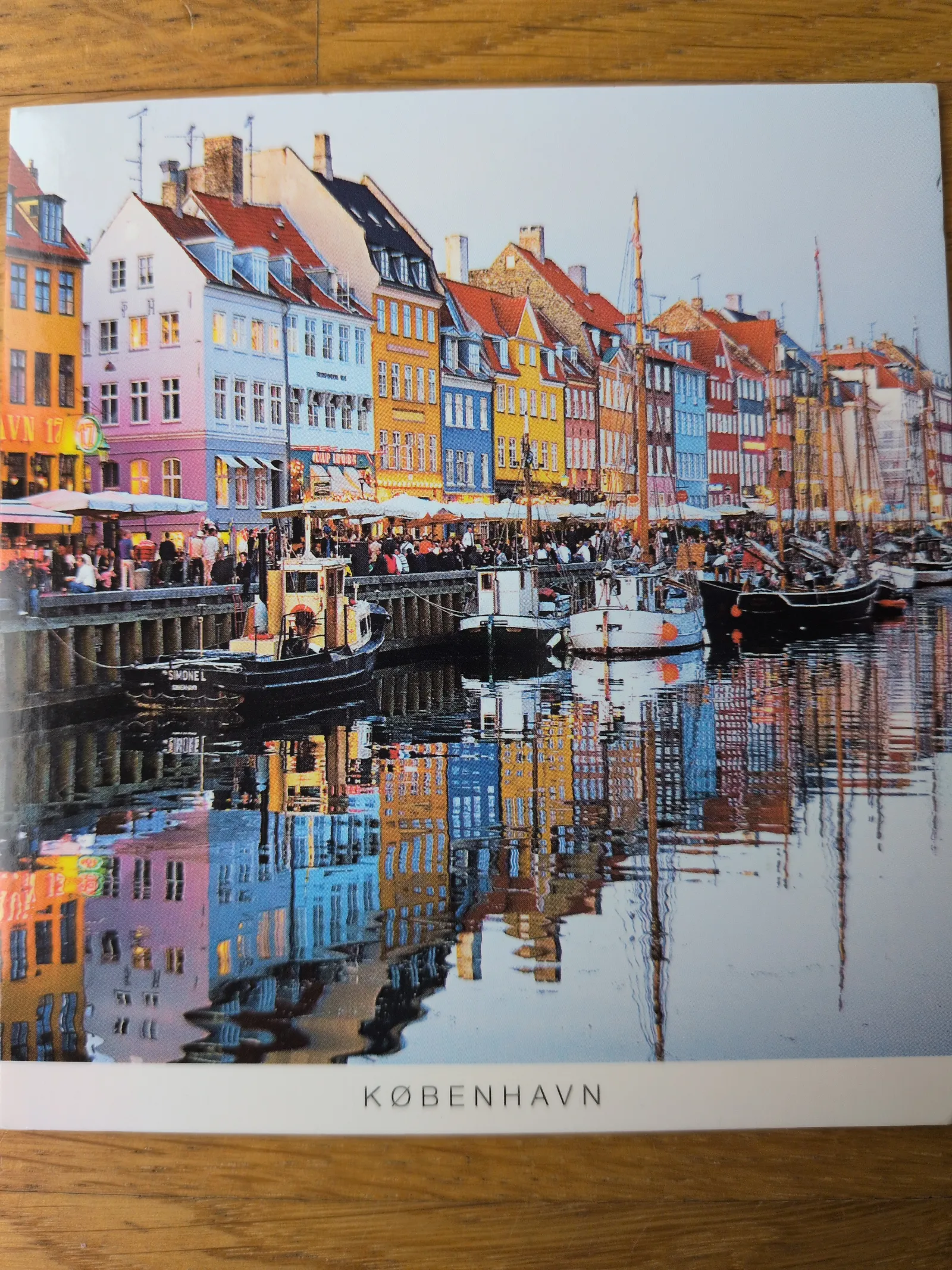 Der Nyhavn in Kopenhagen zeigt sich auf diesem farbenfrohen Bild mit bunten Häusern, zahlreichen Booten und Menschen am Kanal, die sich im Wasser spiegeln.
