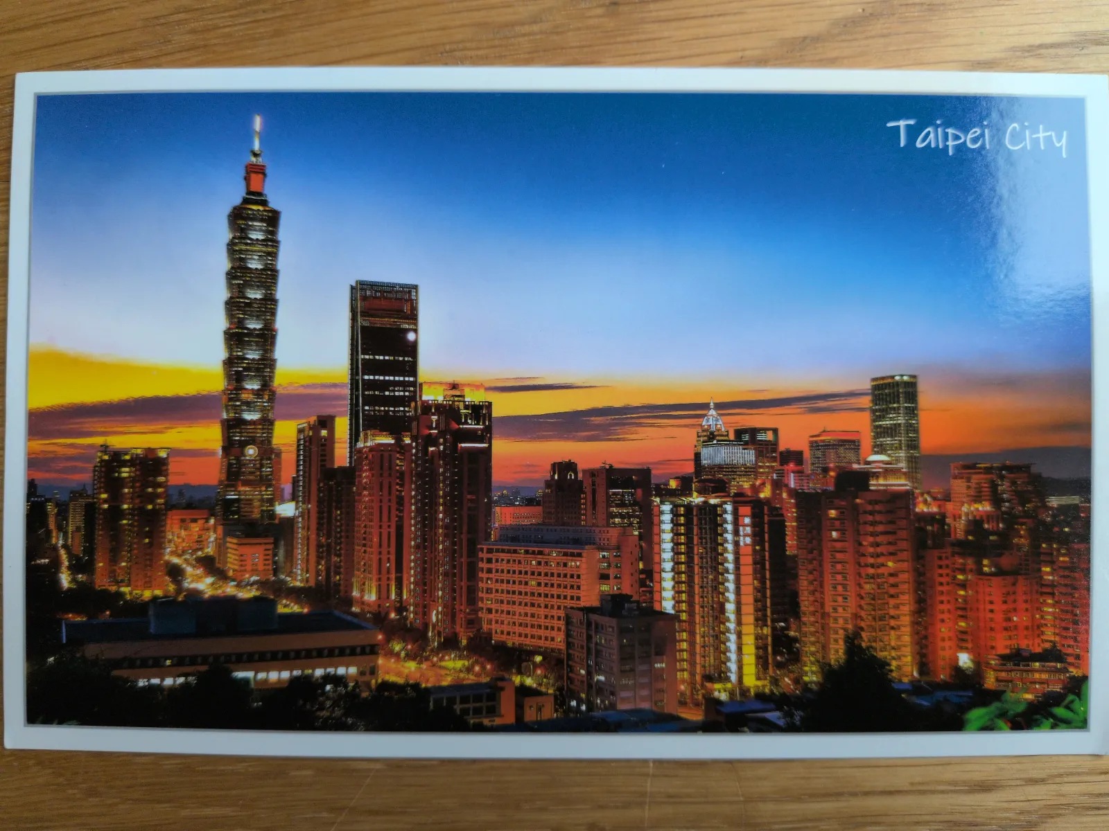 Das Bild zeigt die beleuchtete Skyline von Taipei City bei Sonnenuntergang, mit dem markanten Taipei 101 Hochhaus auf der linken Seite und einem farbenprächtigen Himmel.