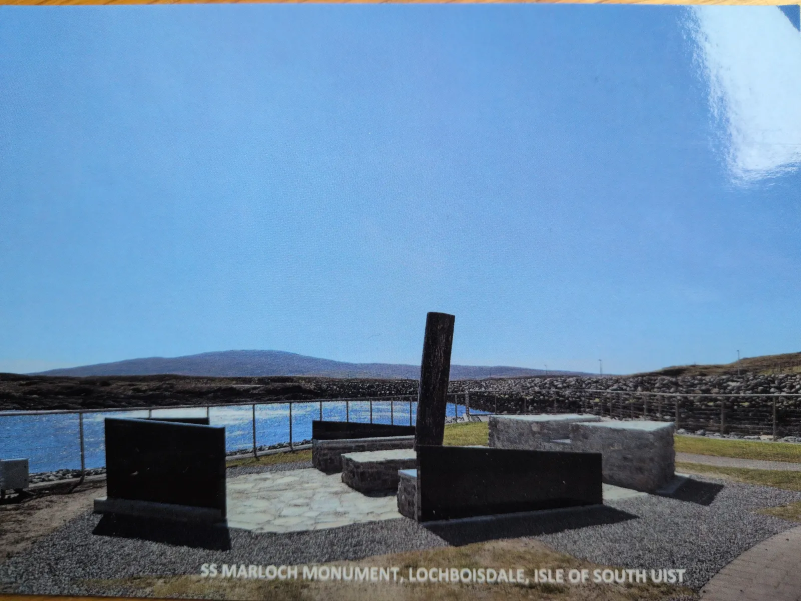 Das SS Marloch Denkmal, eine moderne Skulptur aus dunklen Platten und einem schrägen Pfeiler, steht an einem Seeufer vor einer bergigen Landschaft unter blauem Himmel.