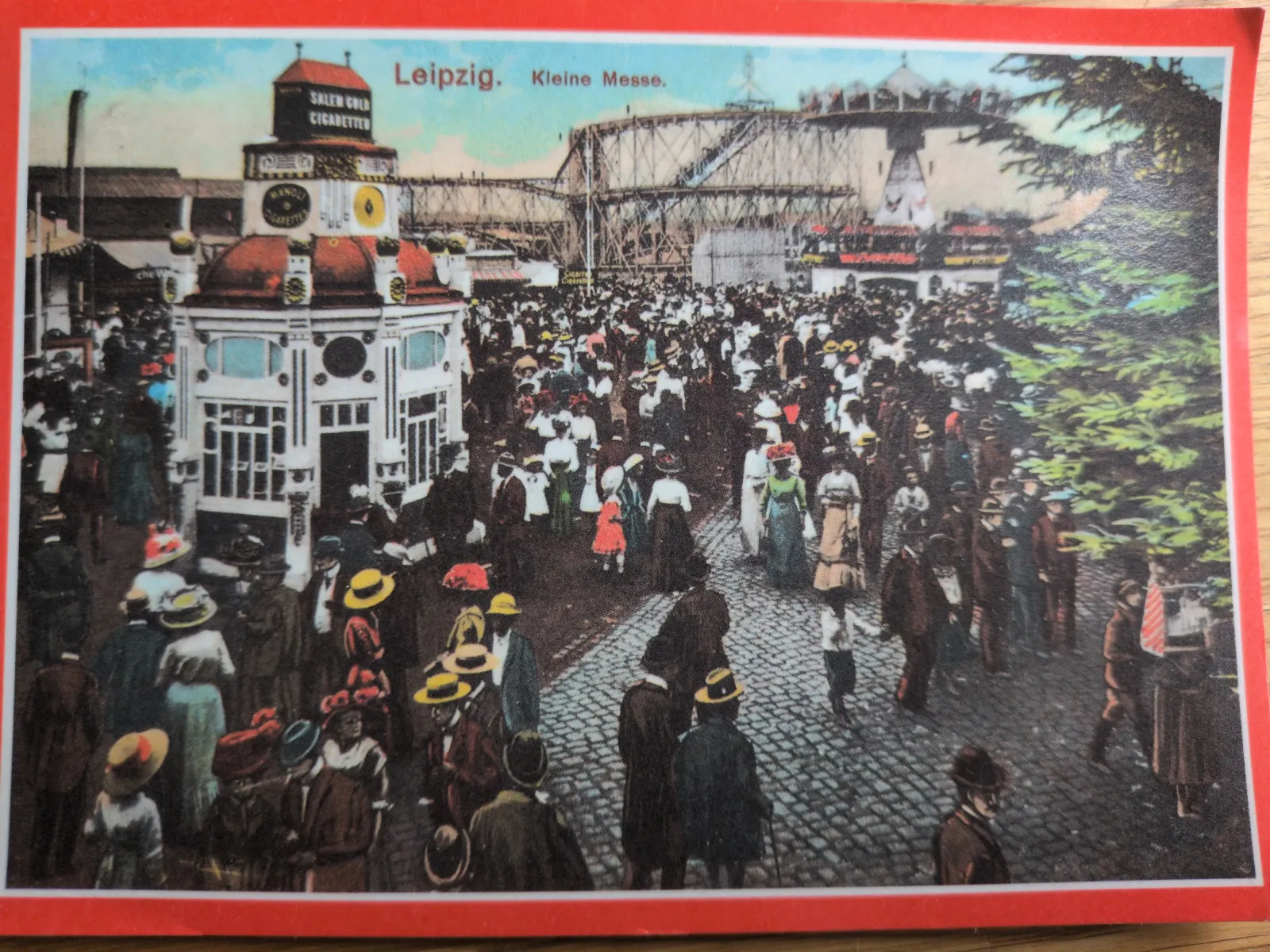 Das Bild zeigt eine historische Szene der "Kleinen Messe" in Leipzig mit einer großen Menschenmenge, einem festlichen Pavillon und einer Achterbahn im Hintergrund.