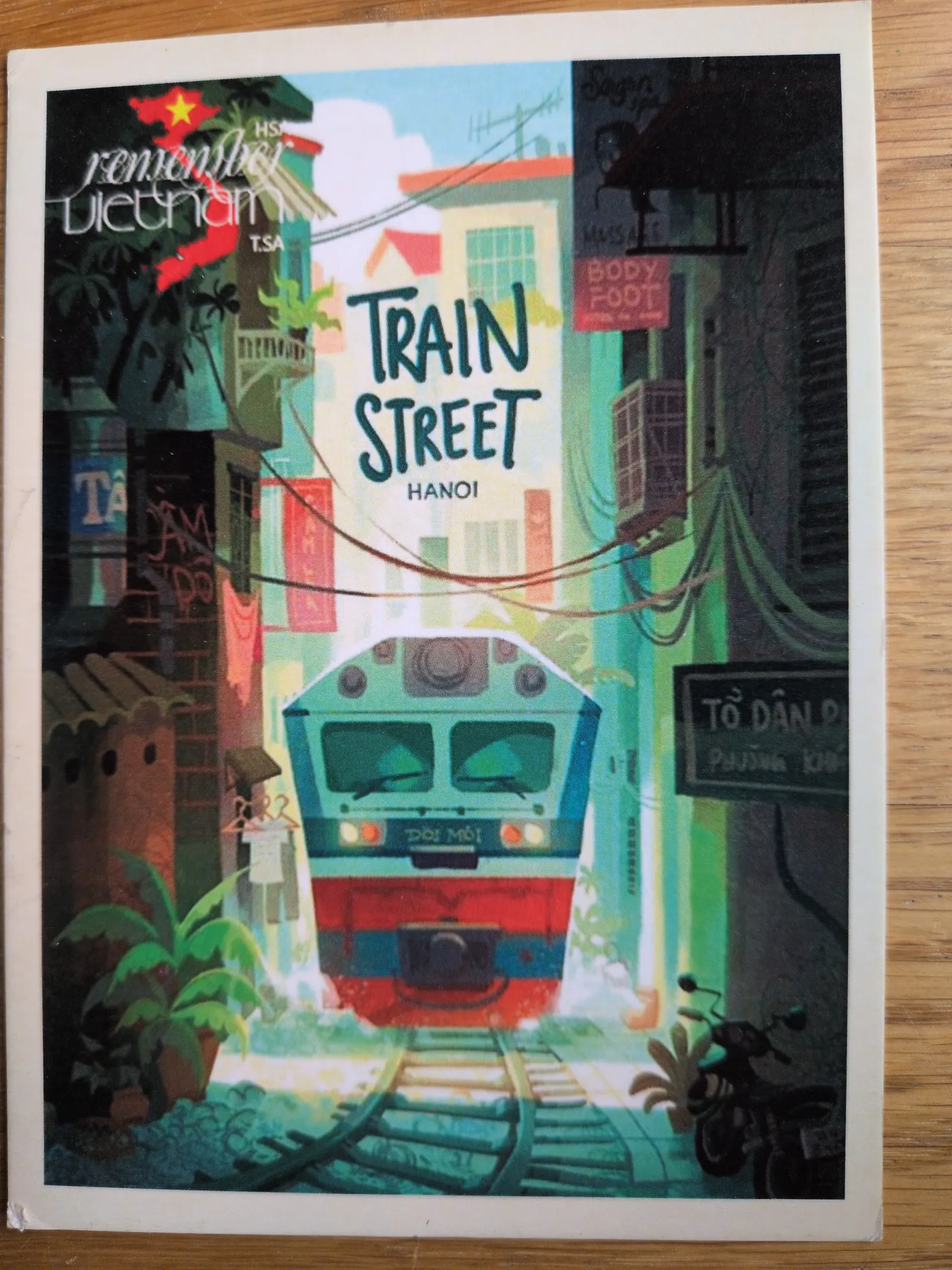 Eine farbige Illustration zeigt einen Zug, der auf einer schmalen Gleisstraße in Hanoi, Vietnam, auf den Betrachter zufährt, umgeben von dicht stehenden Häusern.